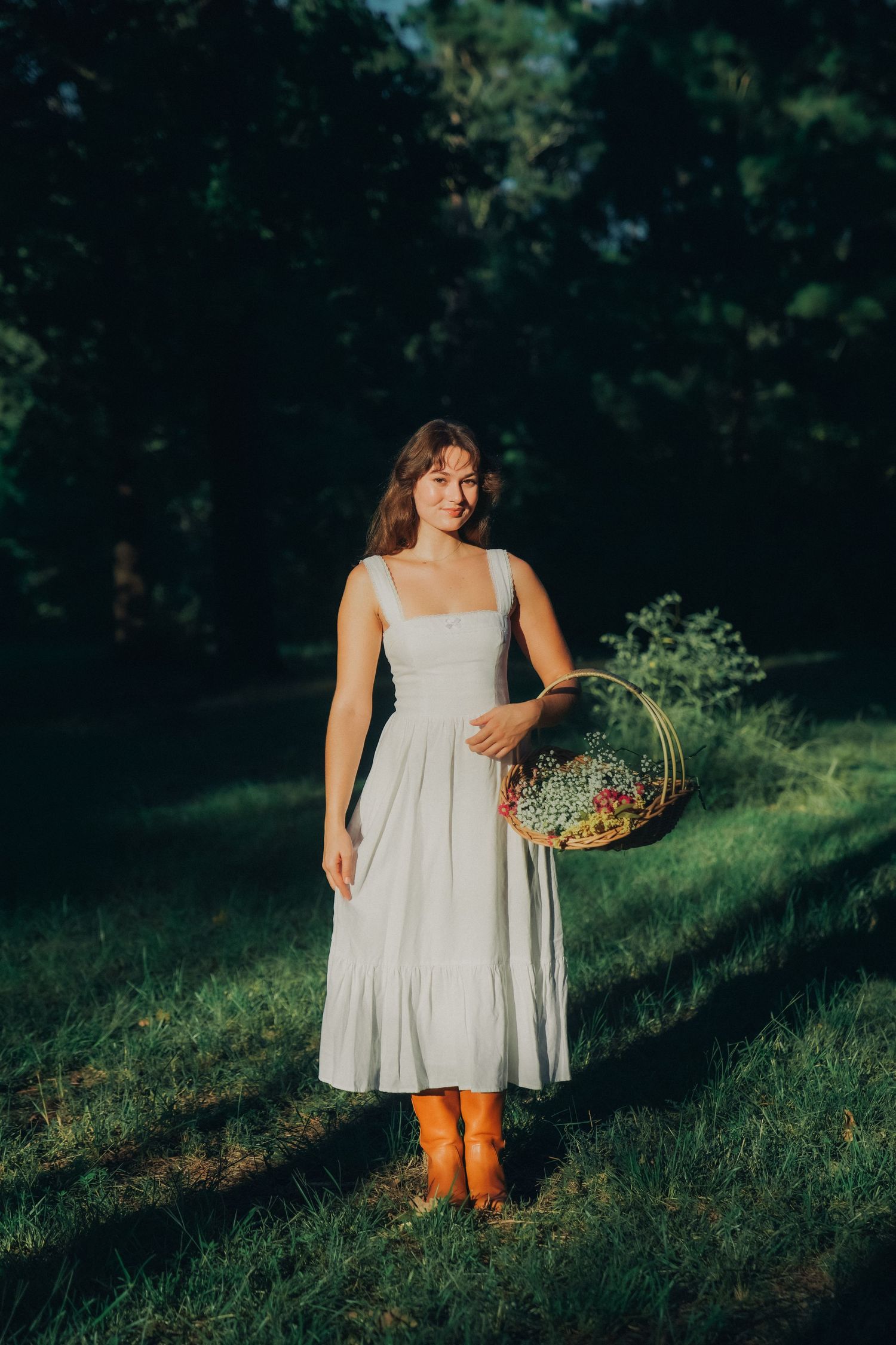 Ethereal scene of a white sundress and boots in a sunlit forest setting with a basket of wildflowers.