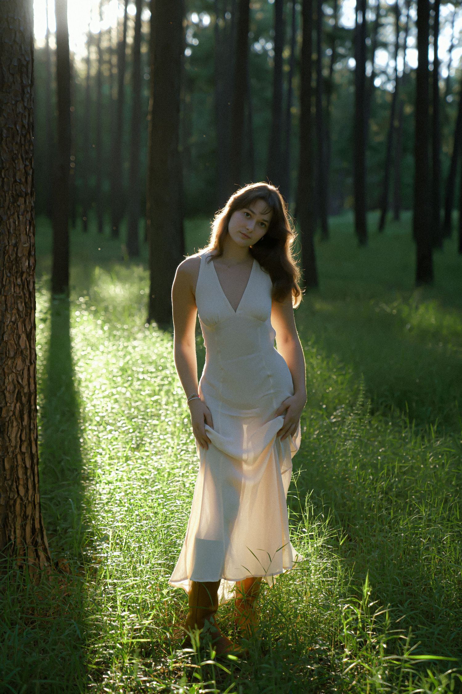 A figure in a flowing white dress and boots stands in a sunlit forest clearing with backlit glow through pine trees.