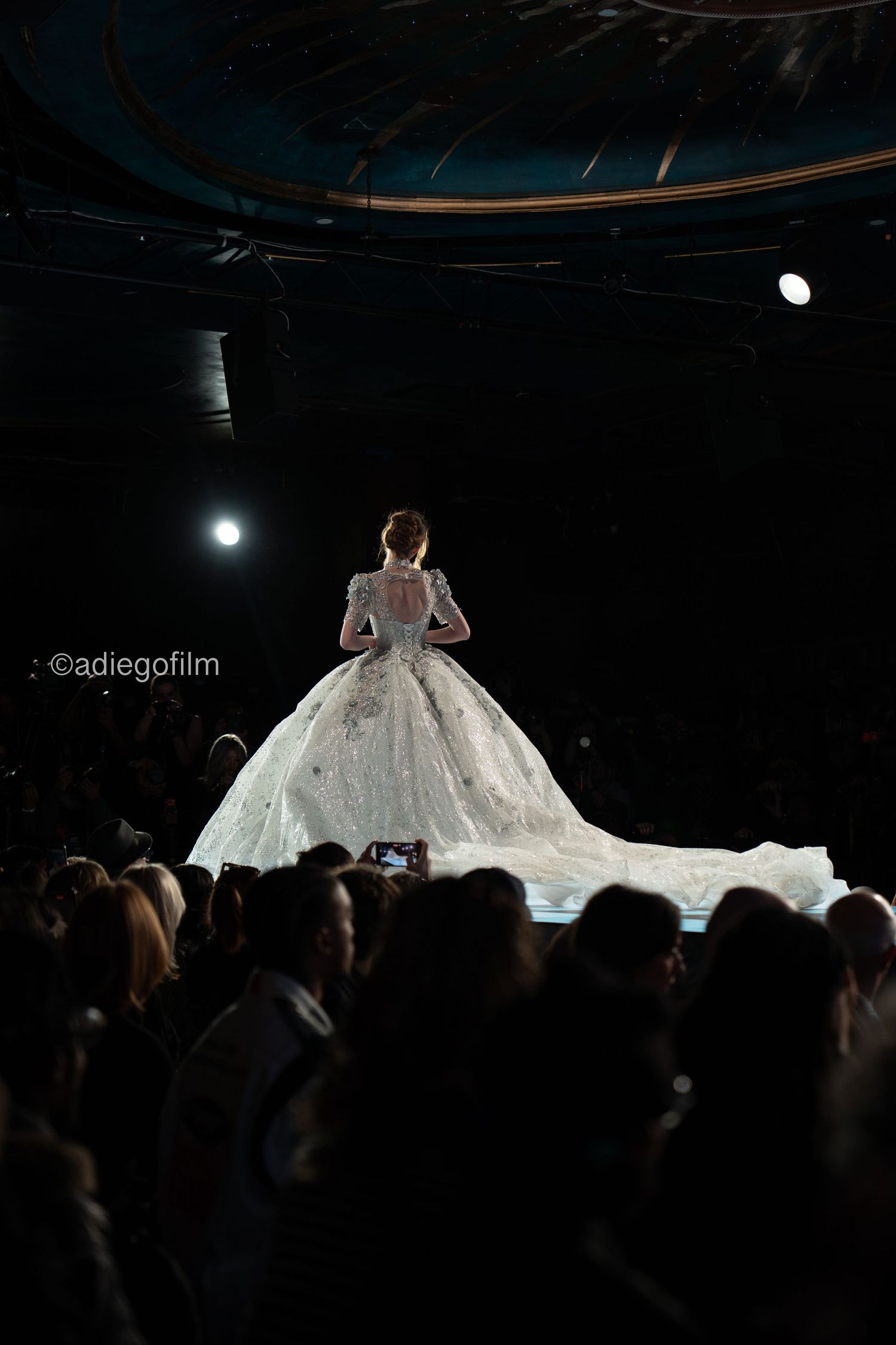 A dramatic fashion show silhouette features an illuminated wedding gown with a flowing train on a dark runway stage.