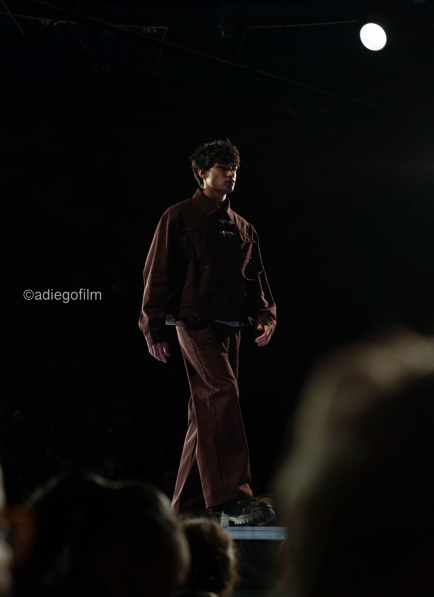 A figure in a brown coat stands in dramatic lighting against a dark background during a stage performance.