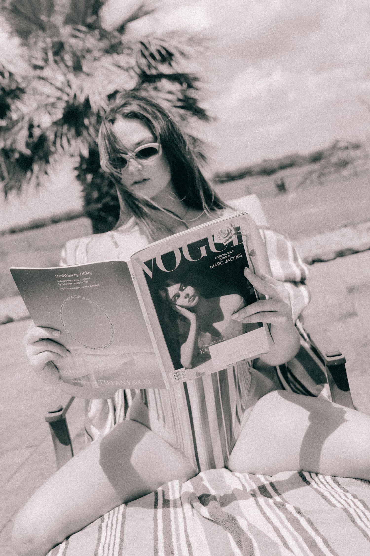 Woman in sunglasses reading Vogue magazine outdoors in retro black and white photography.