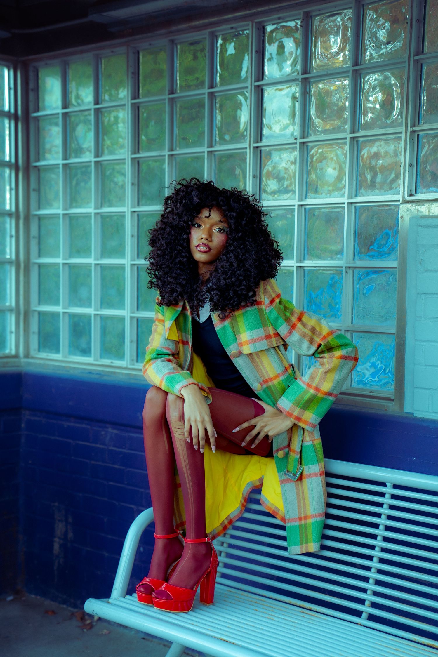 A person in a vibrant plaid jacket poses on a blue bench against a glass block wall with teal lighting.