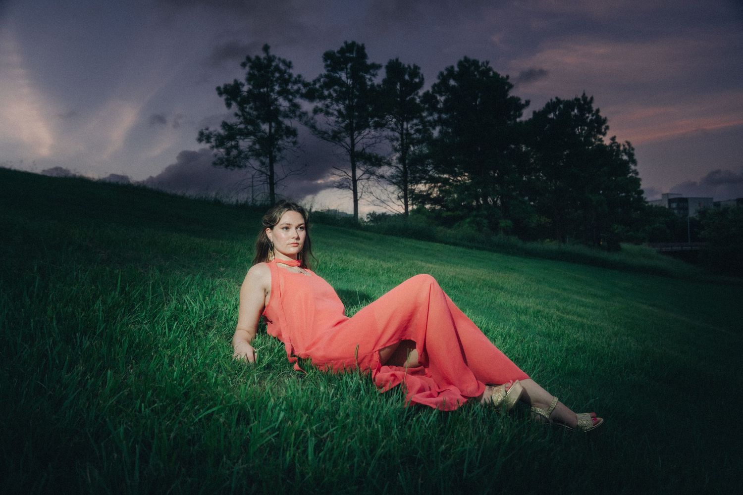 Woman in coral dress reclining on lush green grass at dusk with dramatic purple sky and trees.