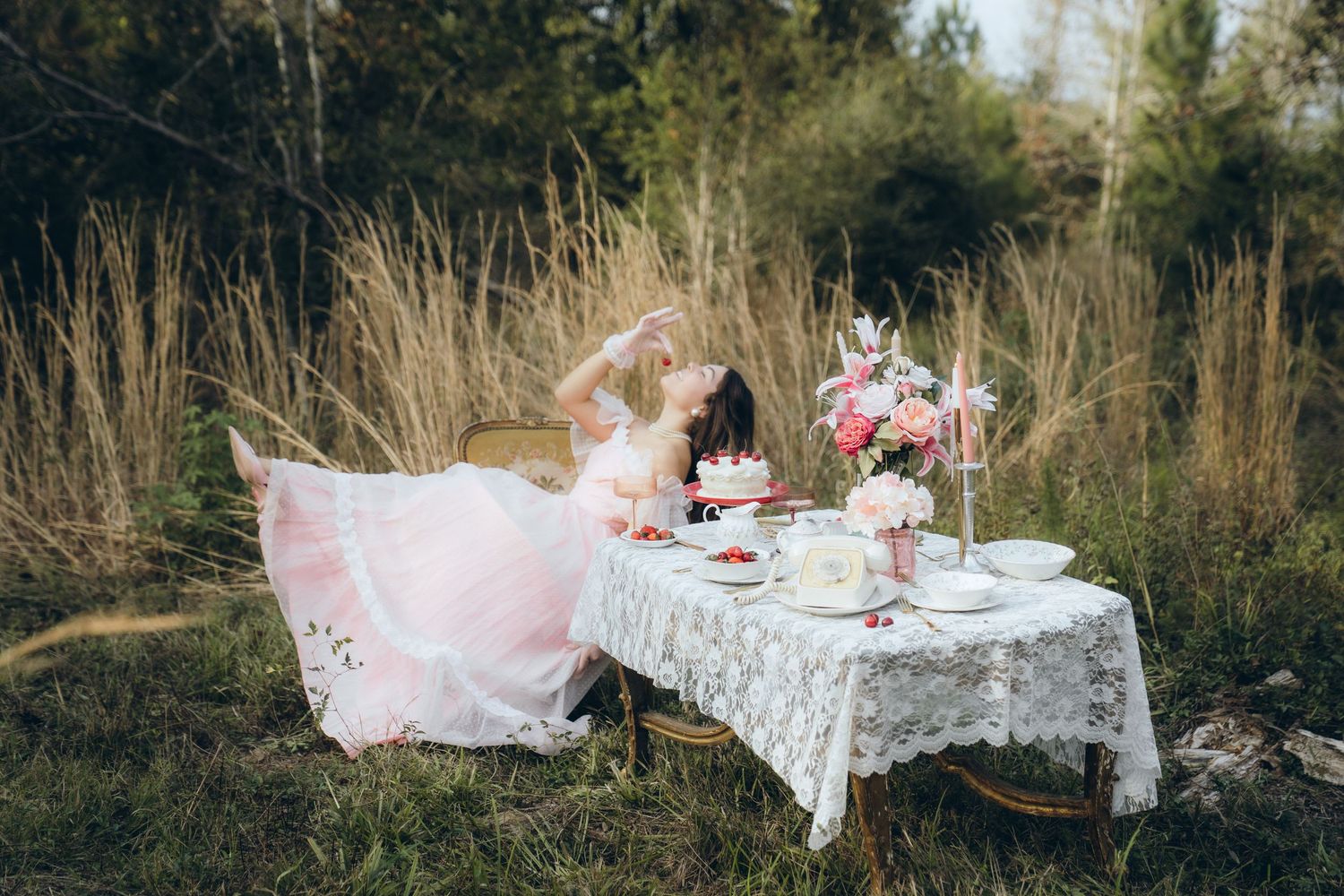 Elegant outdoor tea party setup with white lace tablecloth, pink decor and delicate china in a natural garden setting.