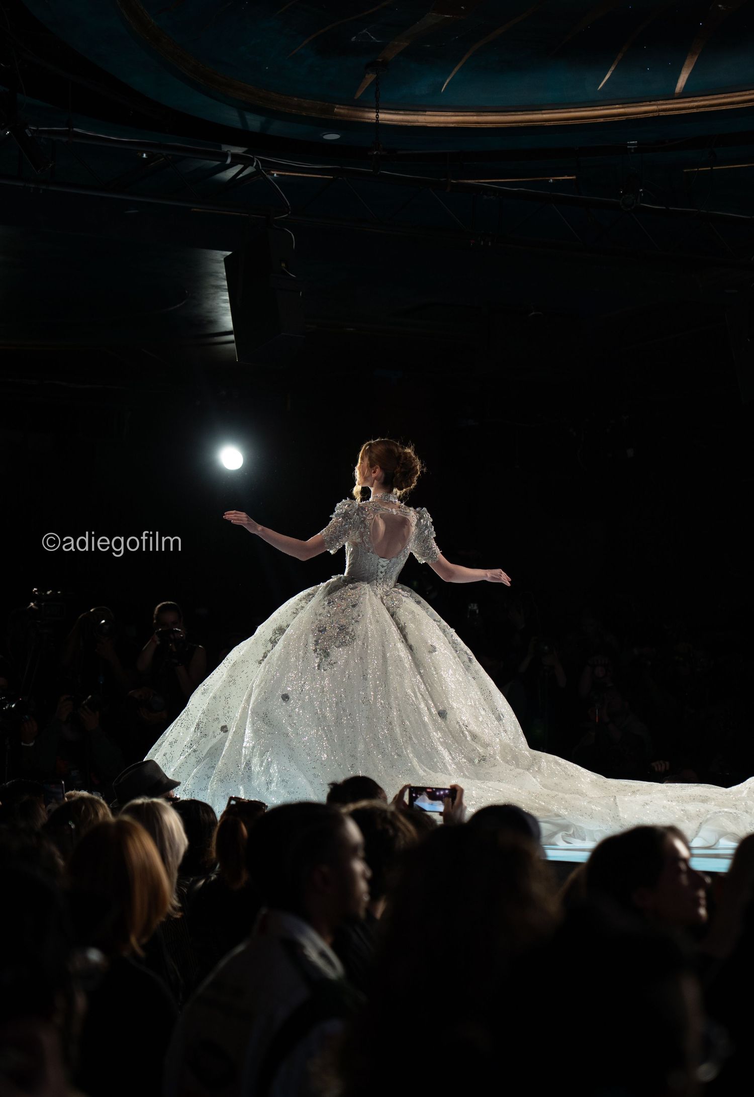 A performer in a flowing white wedding dress twirls dramatically on stage during a fashion show presentation.