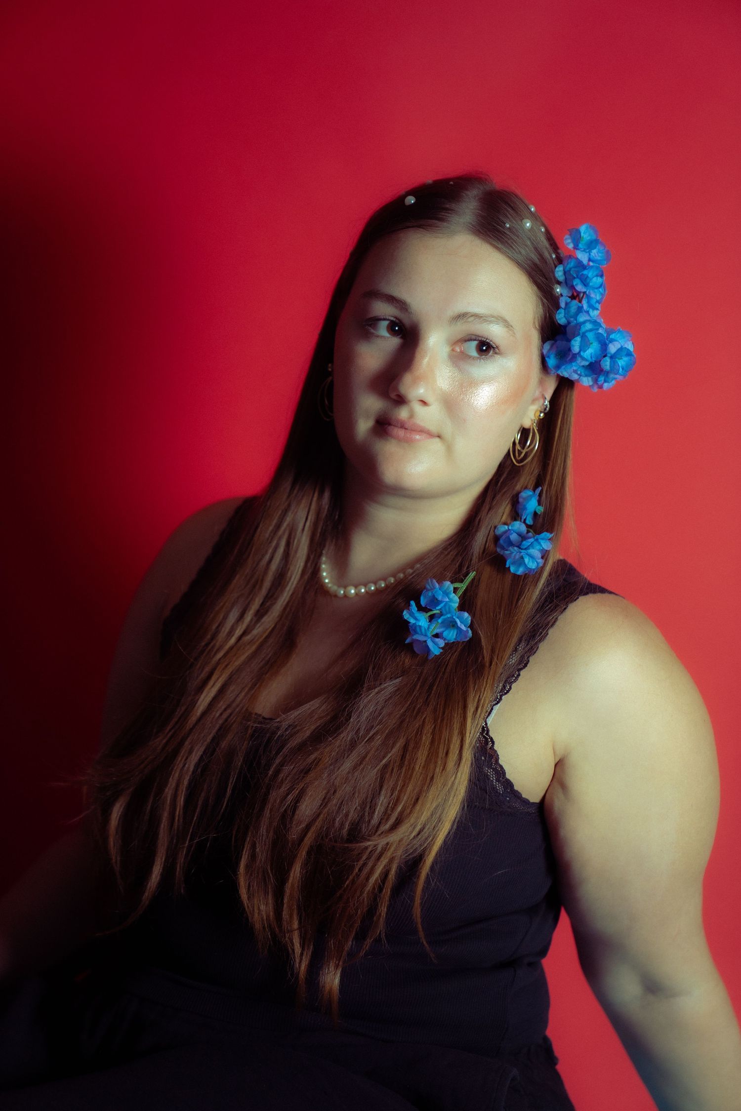 A portrait featuring a person in a dark tank top with bright blue flowers adorning their long brown hair against red backdrop.