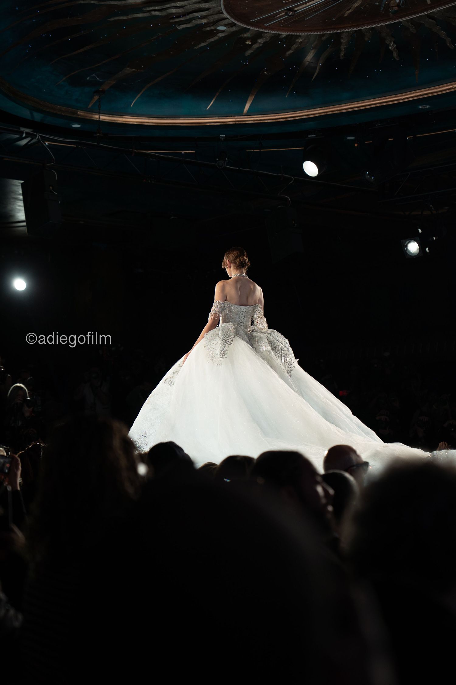 A bride in a voluminous white wedding gown stands on stage during a fashion show with spotlights and silhouetted audience.