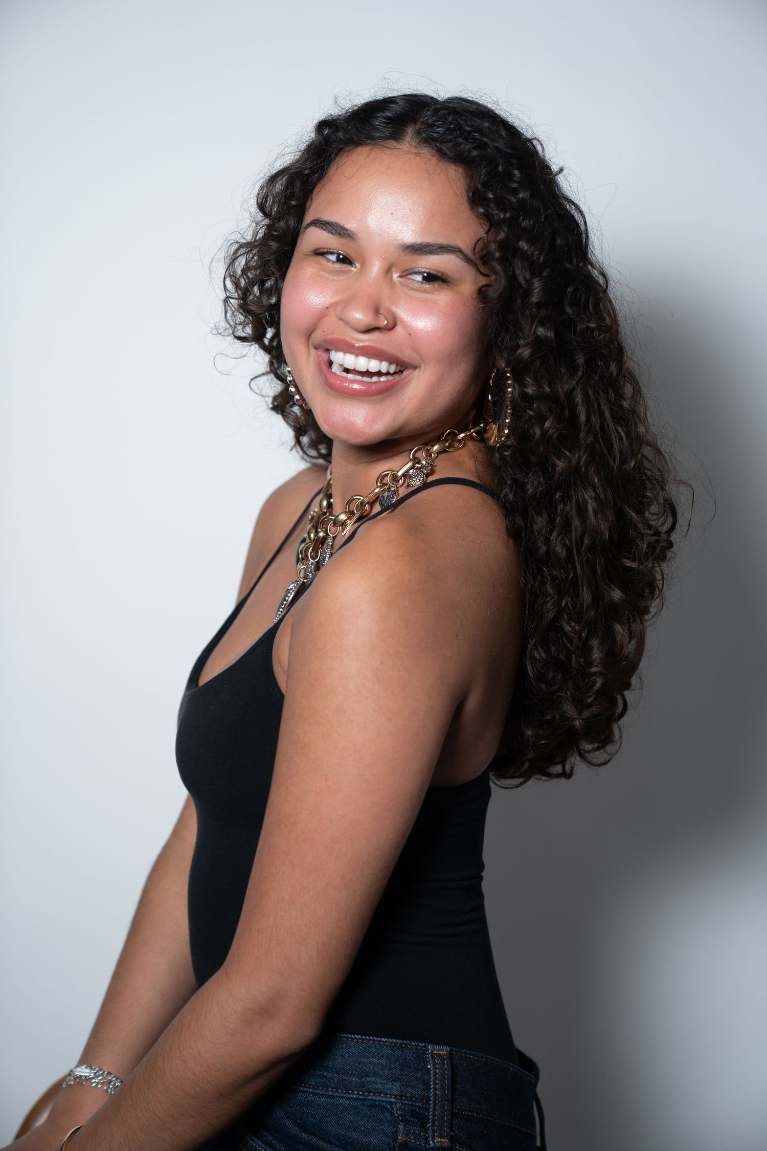 A woman with curly dark hair and a black tank top poses confidently against a light background in a photo series.