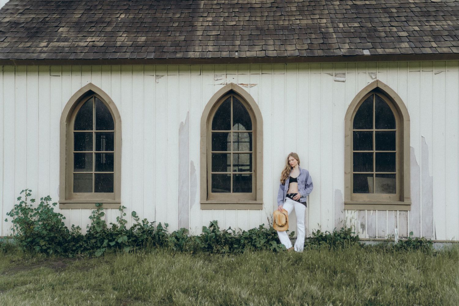 Woman in casual outfit holding a straw hat stands by a white vintage church building with Gothic arch windows.