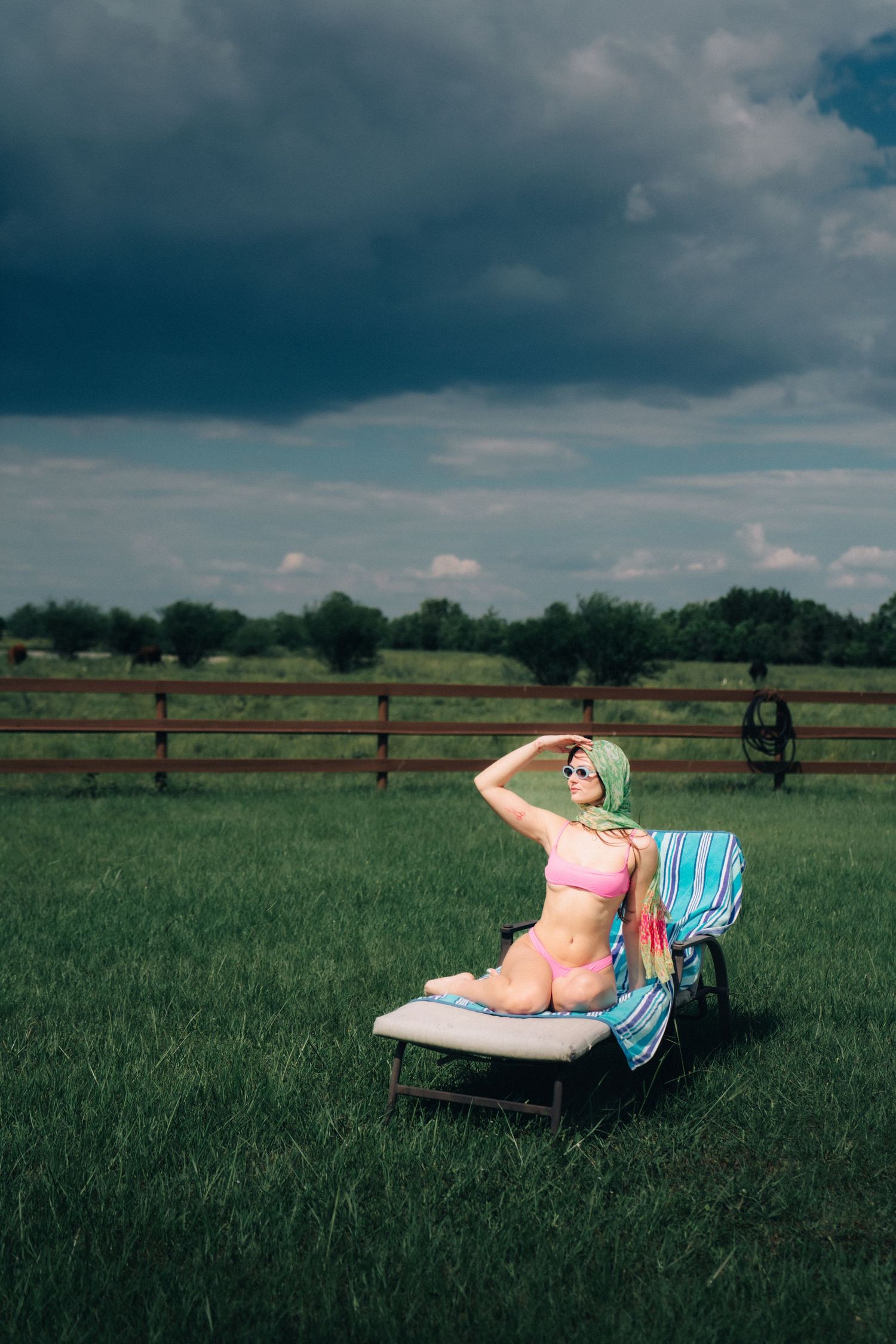 Person lounging on lawn chair in pink swimsuit by wooden fence under dramatic stormy sky.
