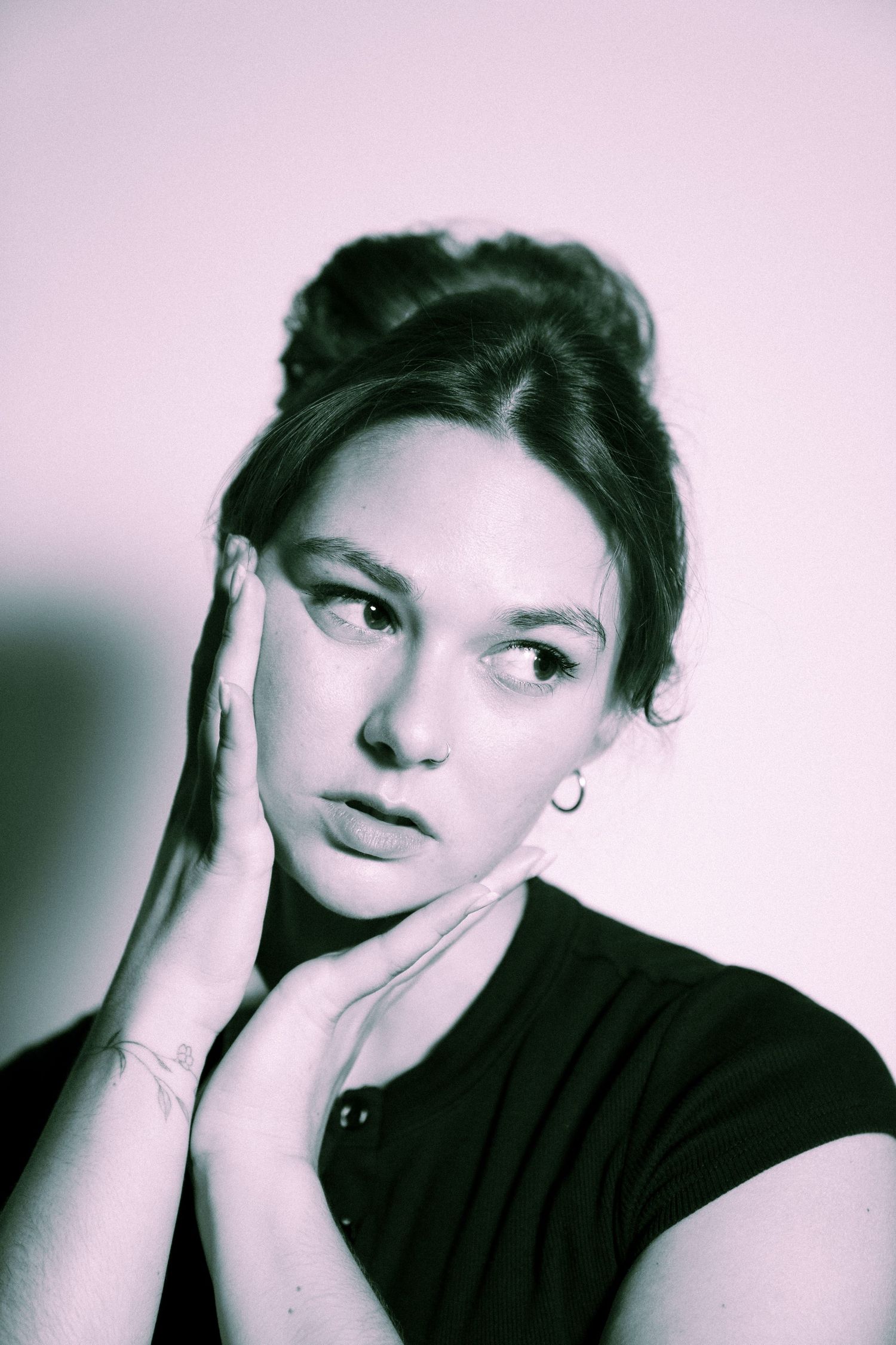 Artistic portrait in black and white with pink tint shows person in dark top with hand resting on their cheek.
