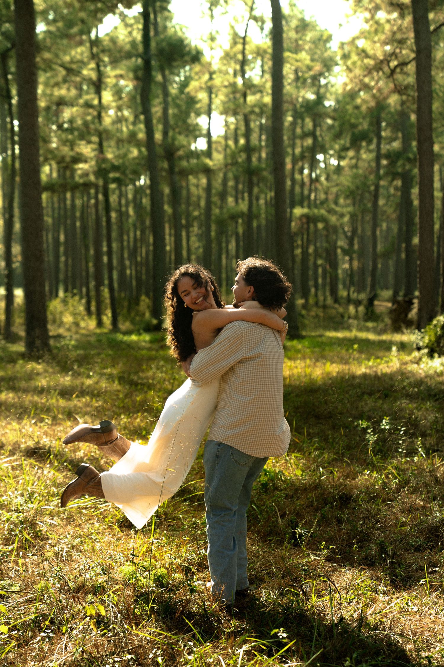 A couple embraces playfully in a sun-dappled forest with golden light filtering through tall pine trees.