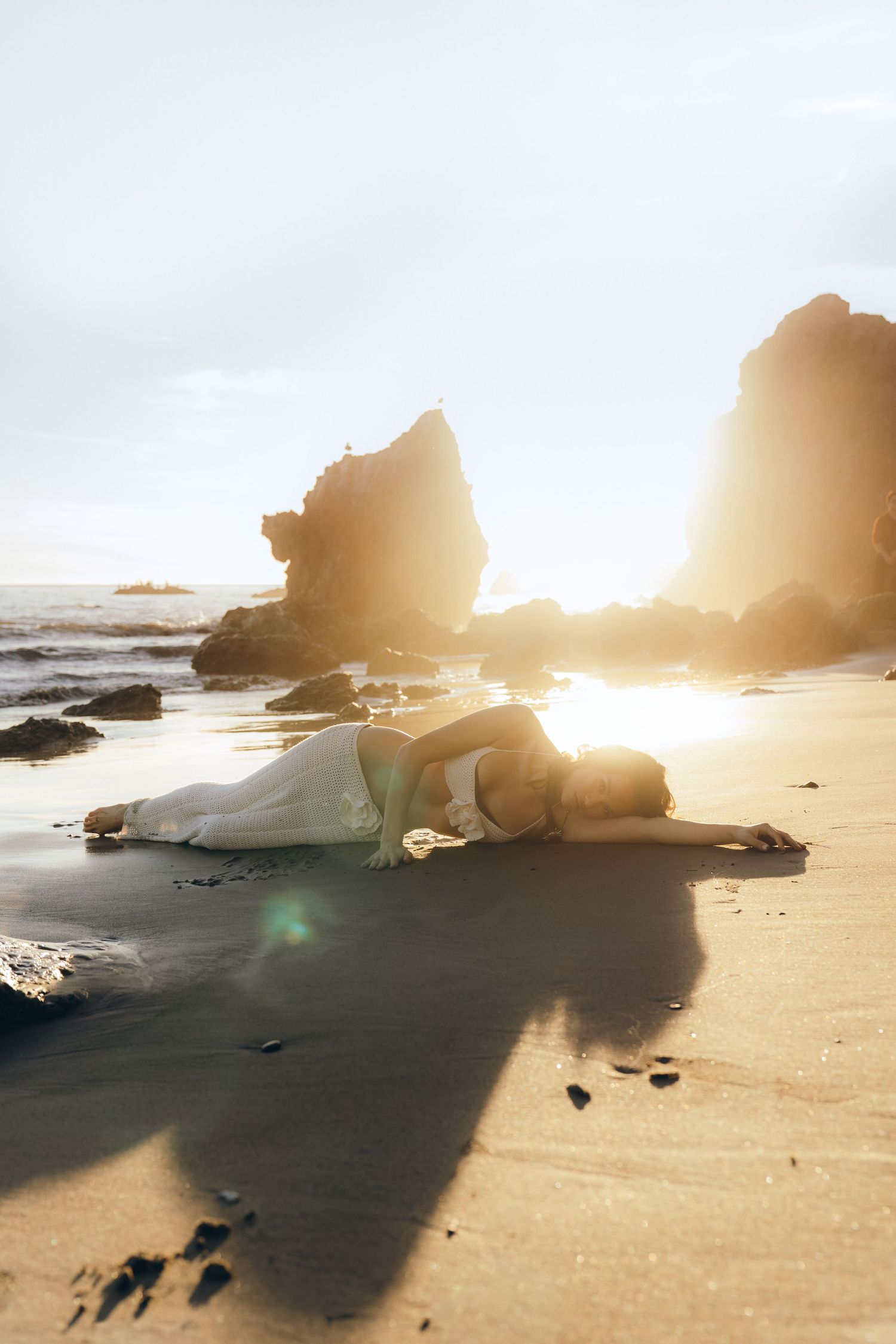 Woman in white dress lying on a sunlit beach with dramatic rock formations and golden sunset light.