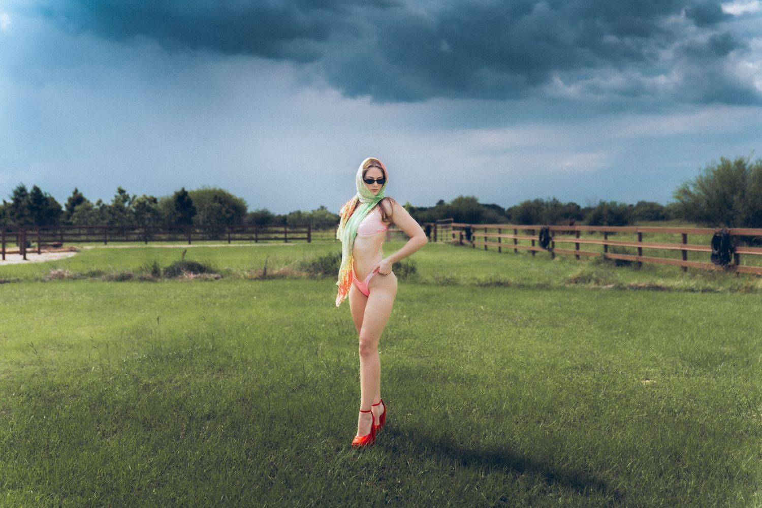 A person poses in a field with storm clouds overhead, wearing colorful clothing and red shoes on a grassy pasture.