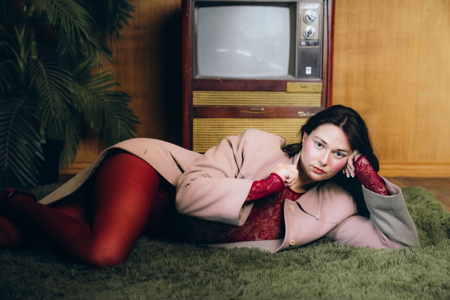 A person in pink clothing reclines on green carpet in front of a wood-paneled wall.