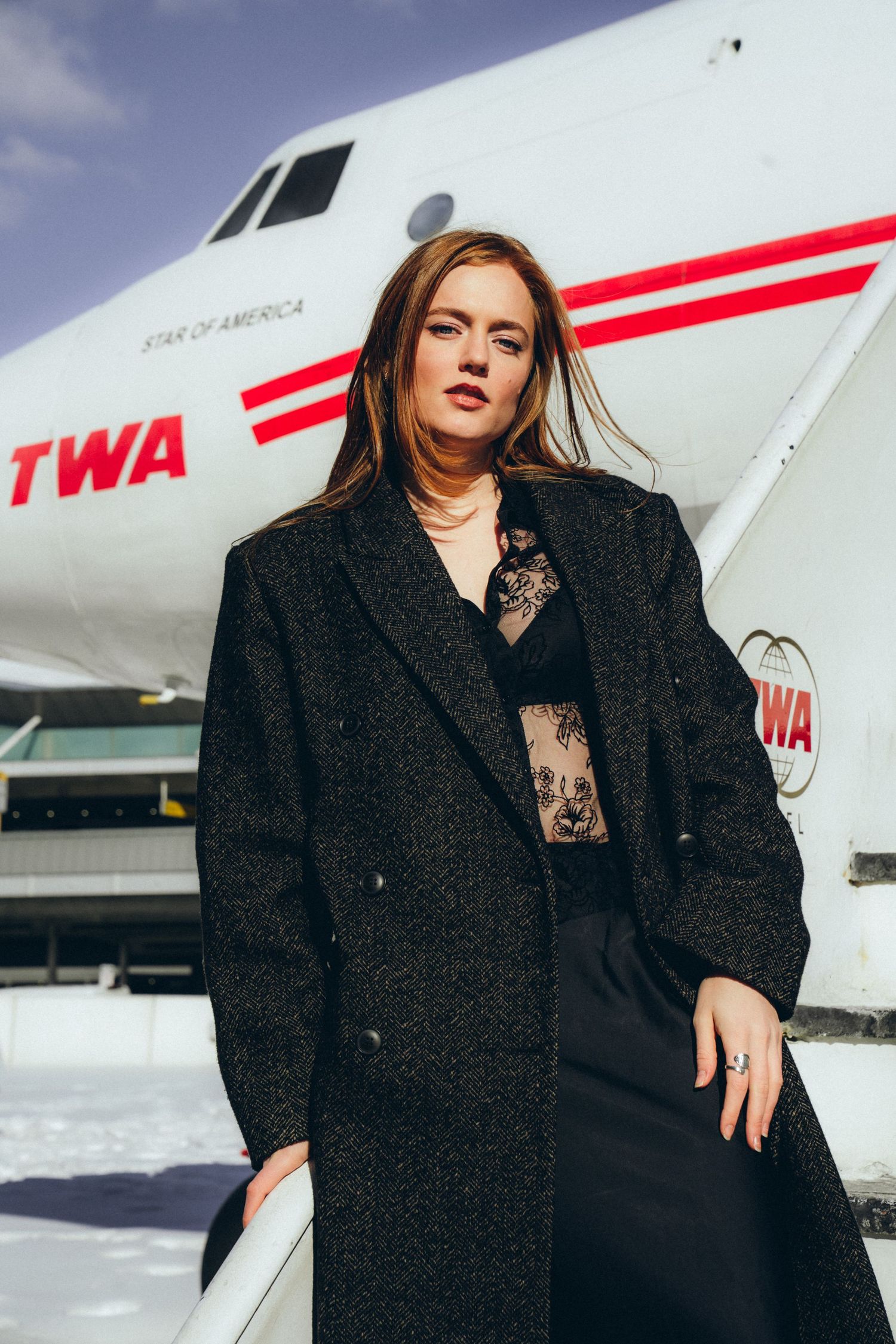 Fashion model in black winter coat poses against vintage TWA aircraft in dramatic outdoor shoot.