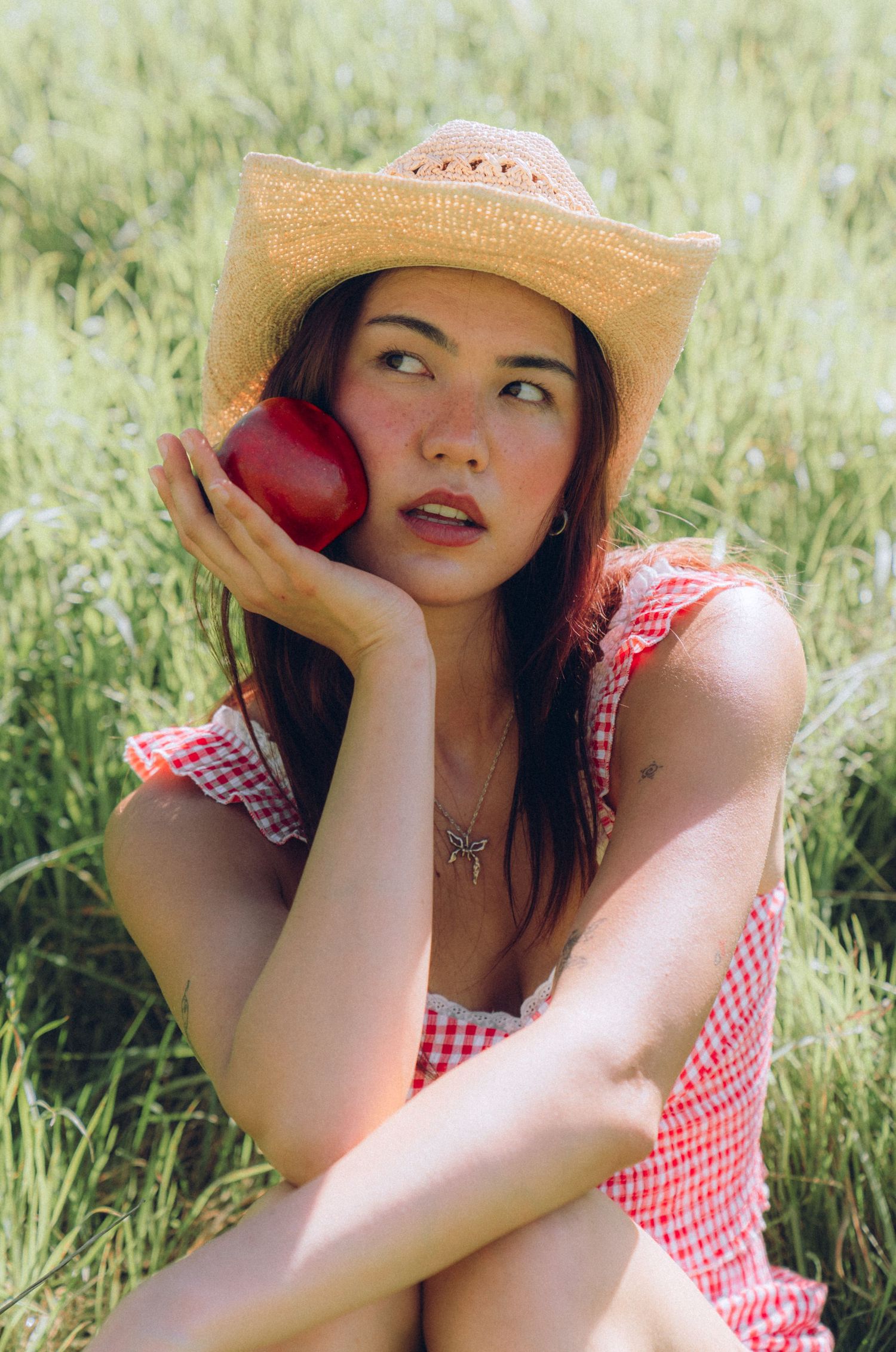 Woman in red gingham dress and straw hat holds red apple while sitting in sunny meadow.