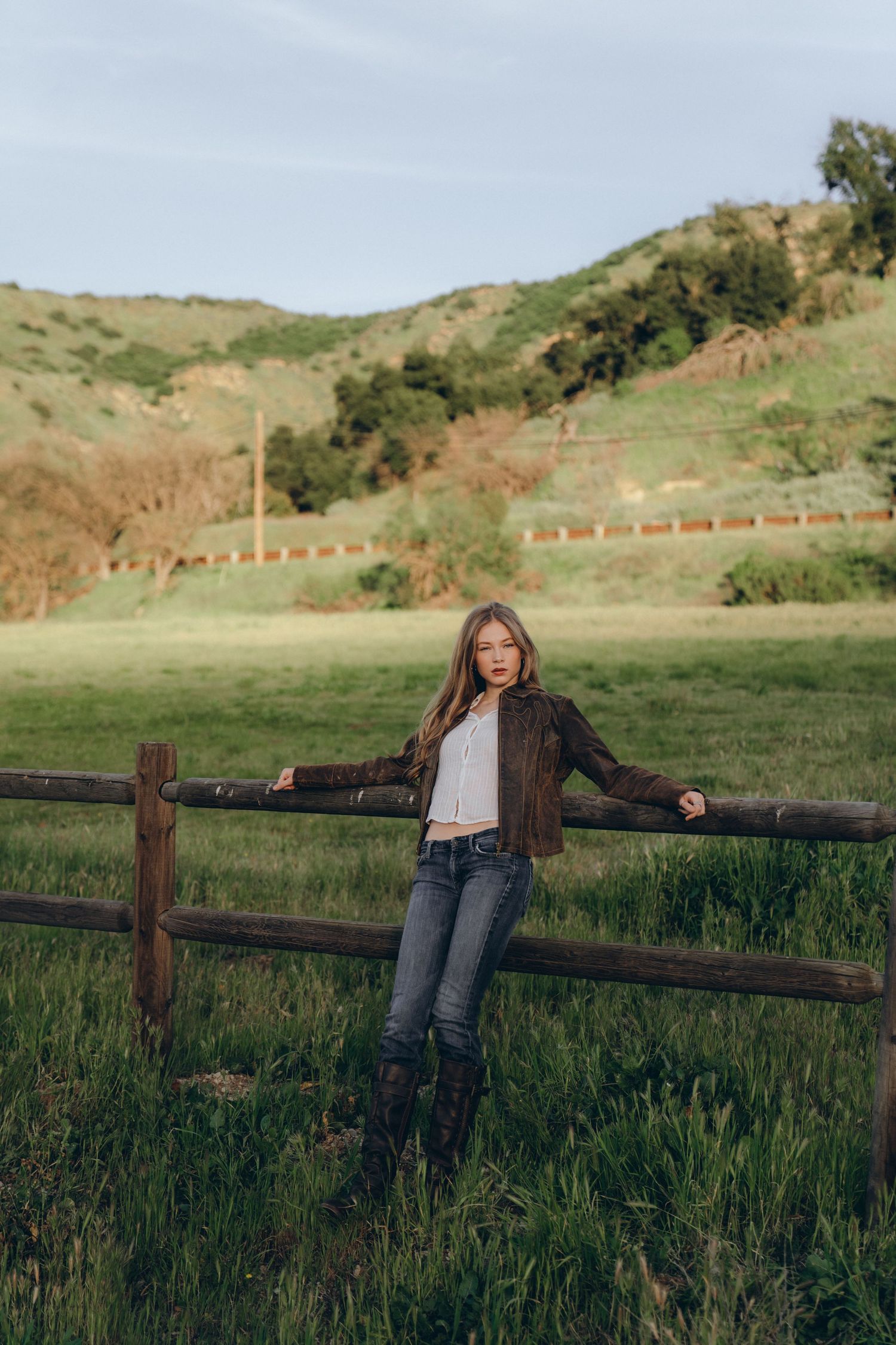 Young woman in brown leather jacket and jeans leaning on rustic wooden fence with green hills in background.