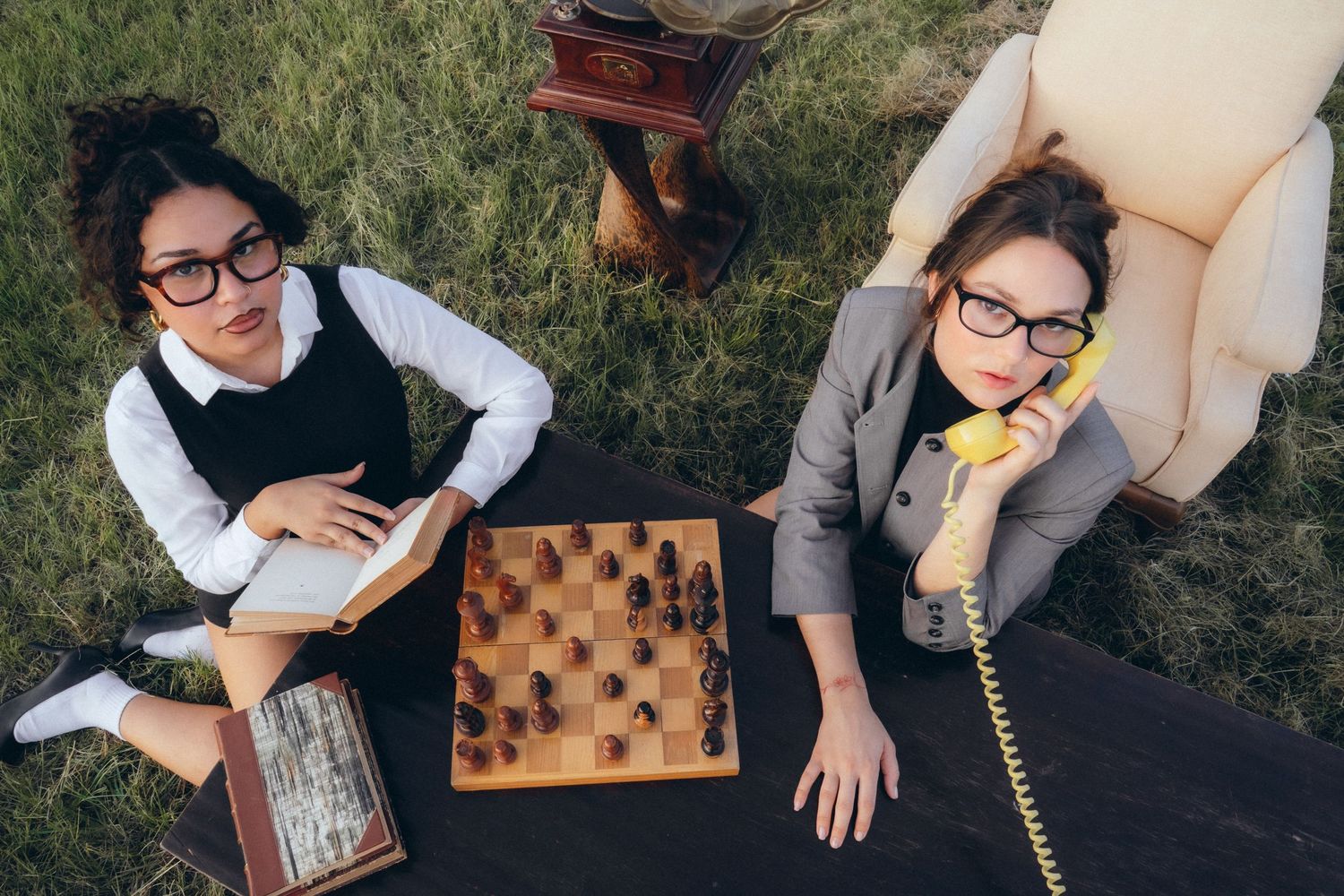 Two people in glasses enjoying a game of chess outdoors while talking on vintage telephones in a quirky scene.