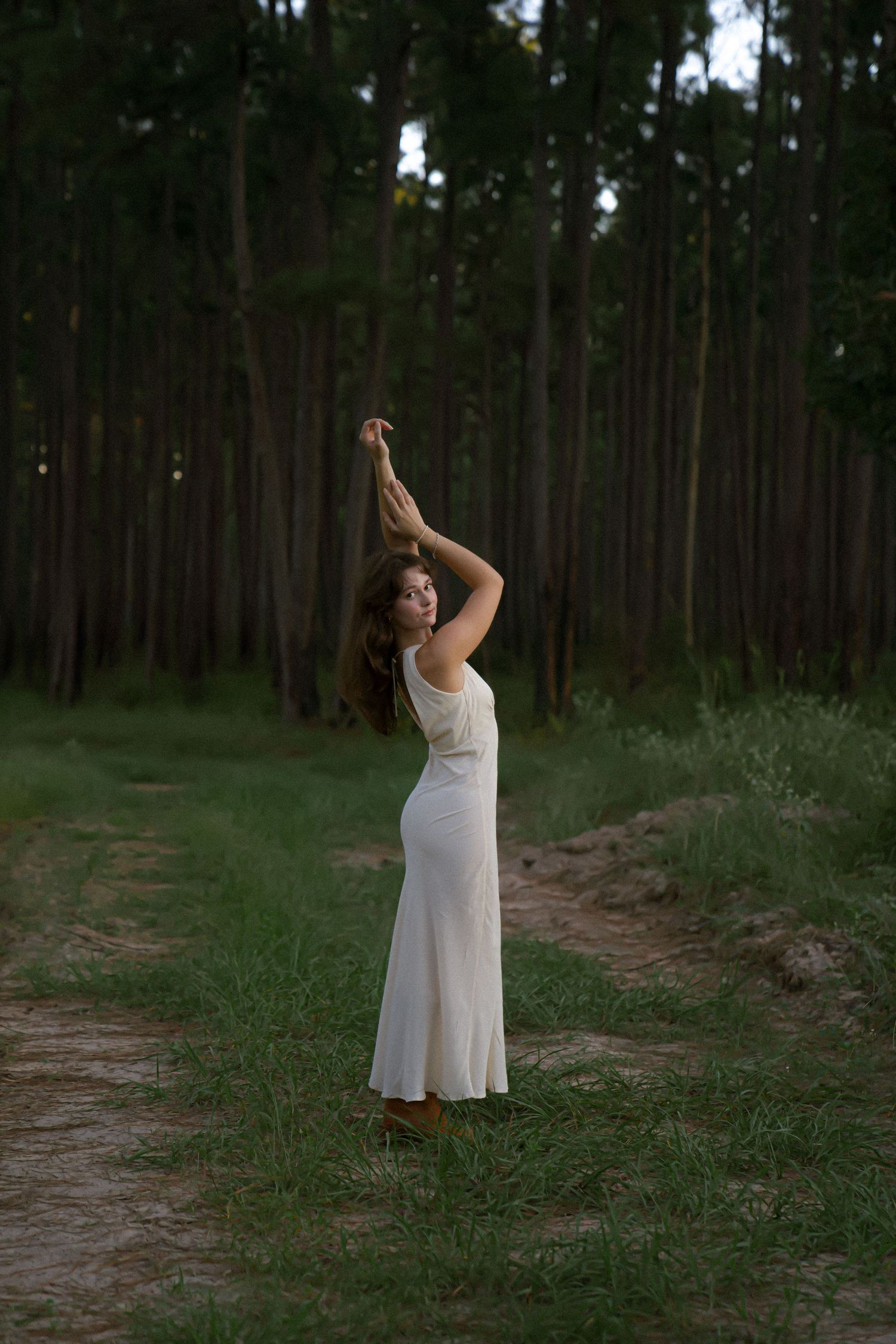 Graceful dance-like pose in white dress on forest path at dusk.