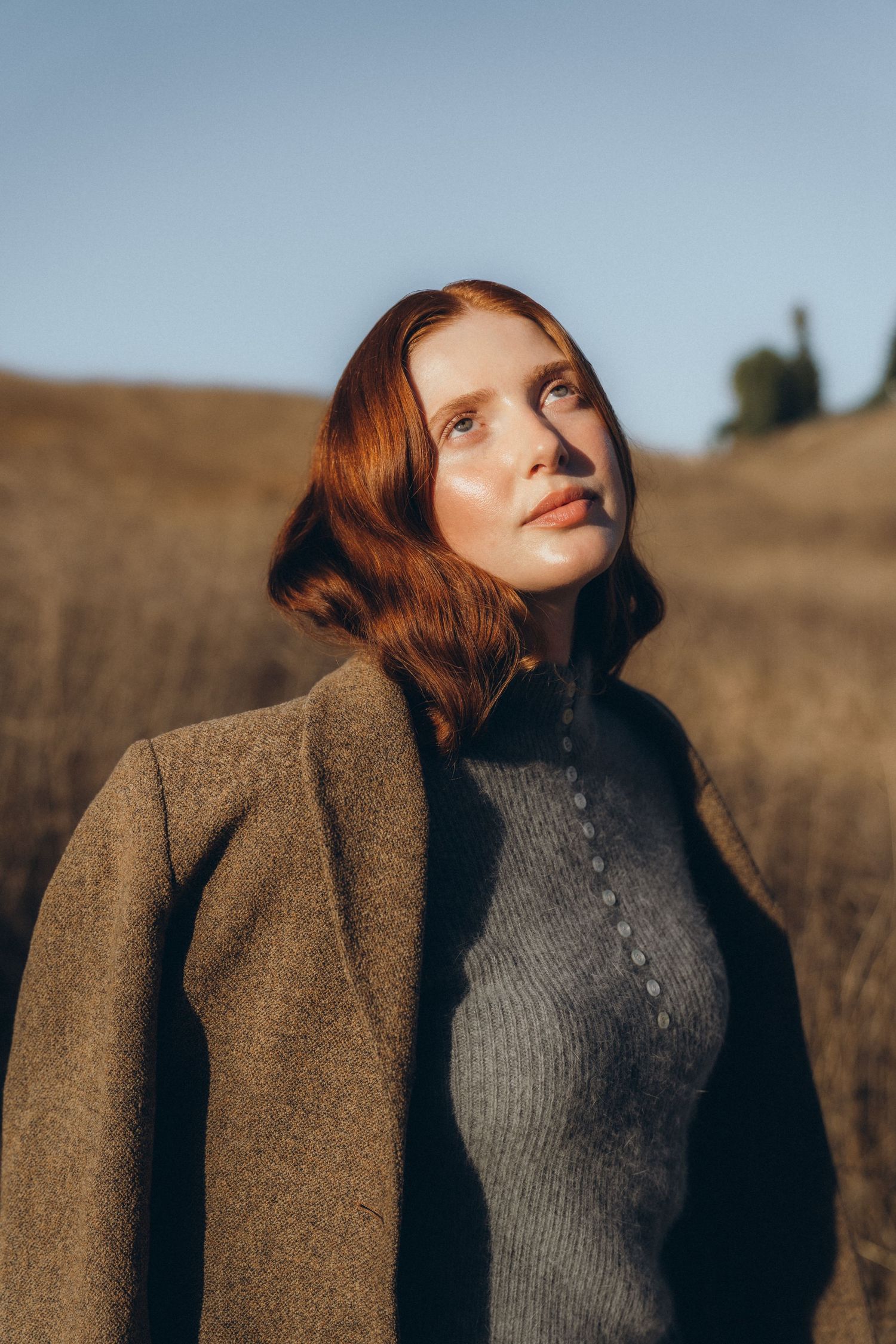 A series of artistic portraits showing someone with red hair wearing a brown coat and gray sweater against a golden field.