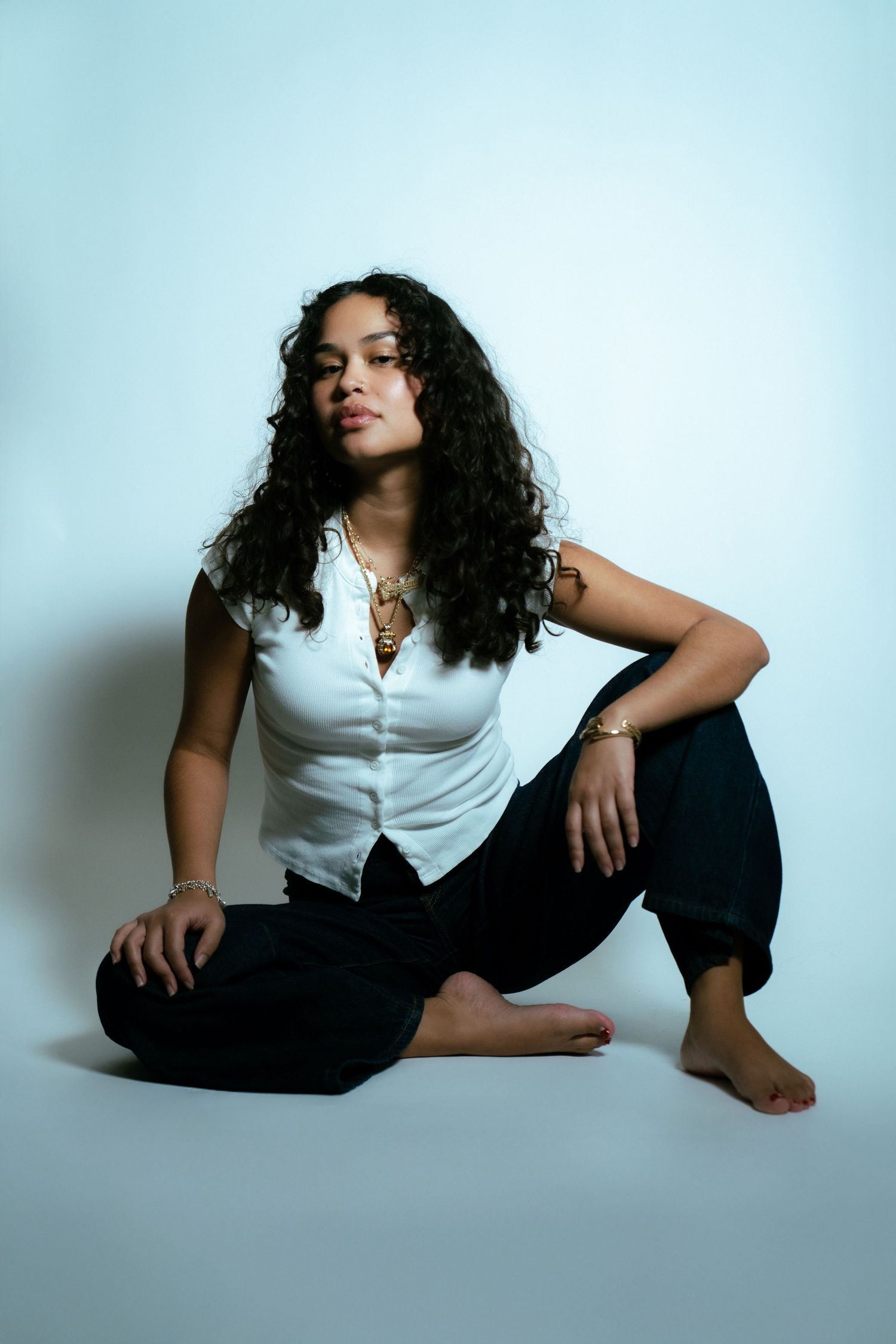 Person with long dark curly hair in white button-up shirt and black pants poses on plain studio background.