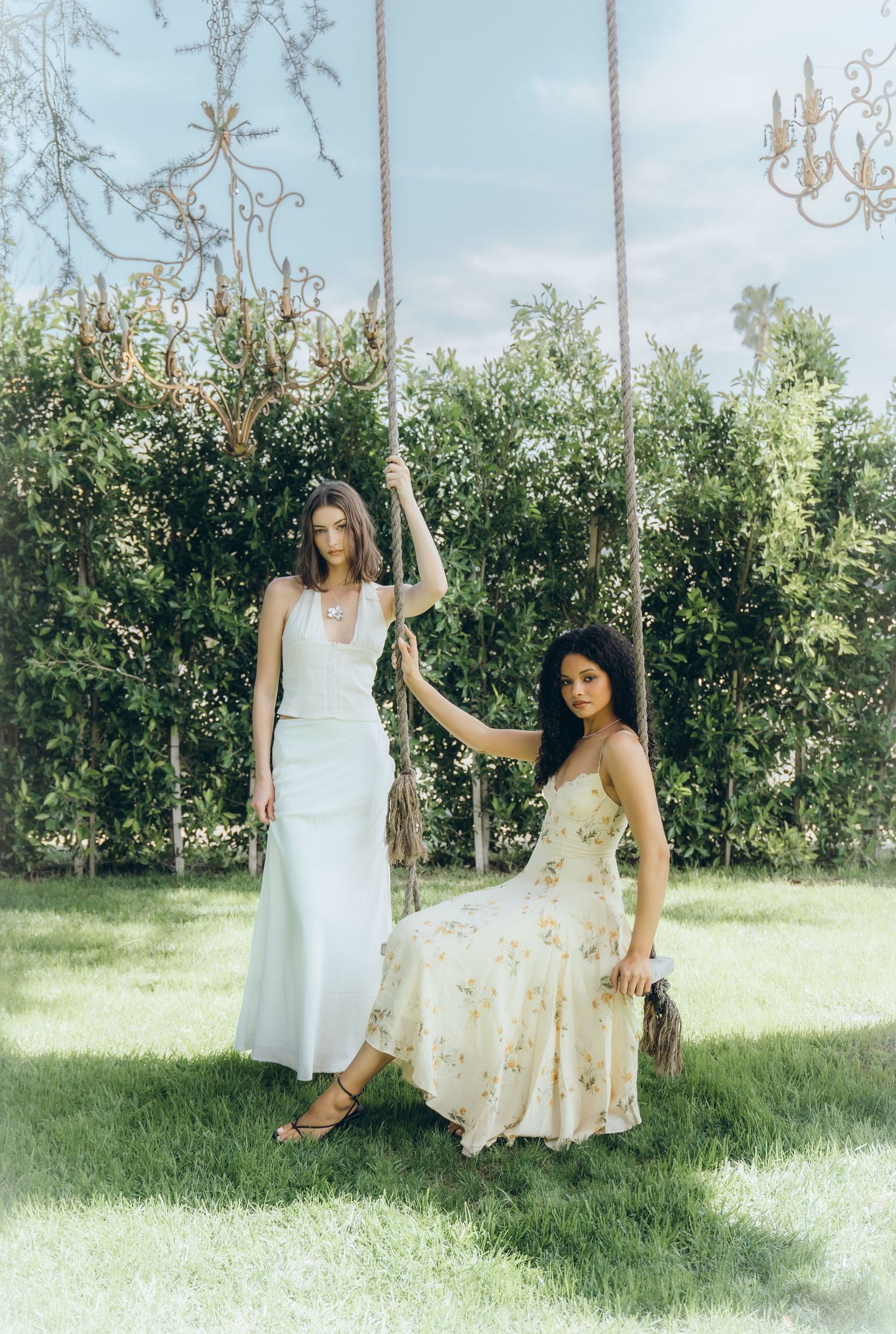 Two women in summer dresses sit and pose together on swings in a sunny garden setting with lush greenery.