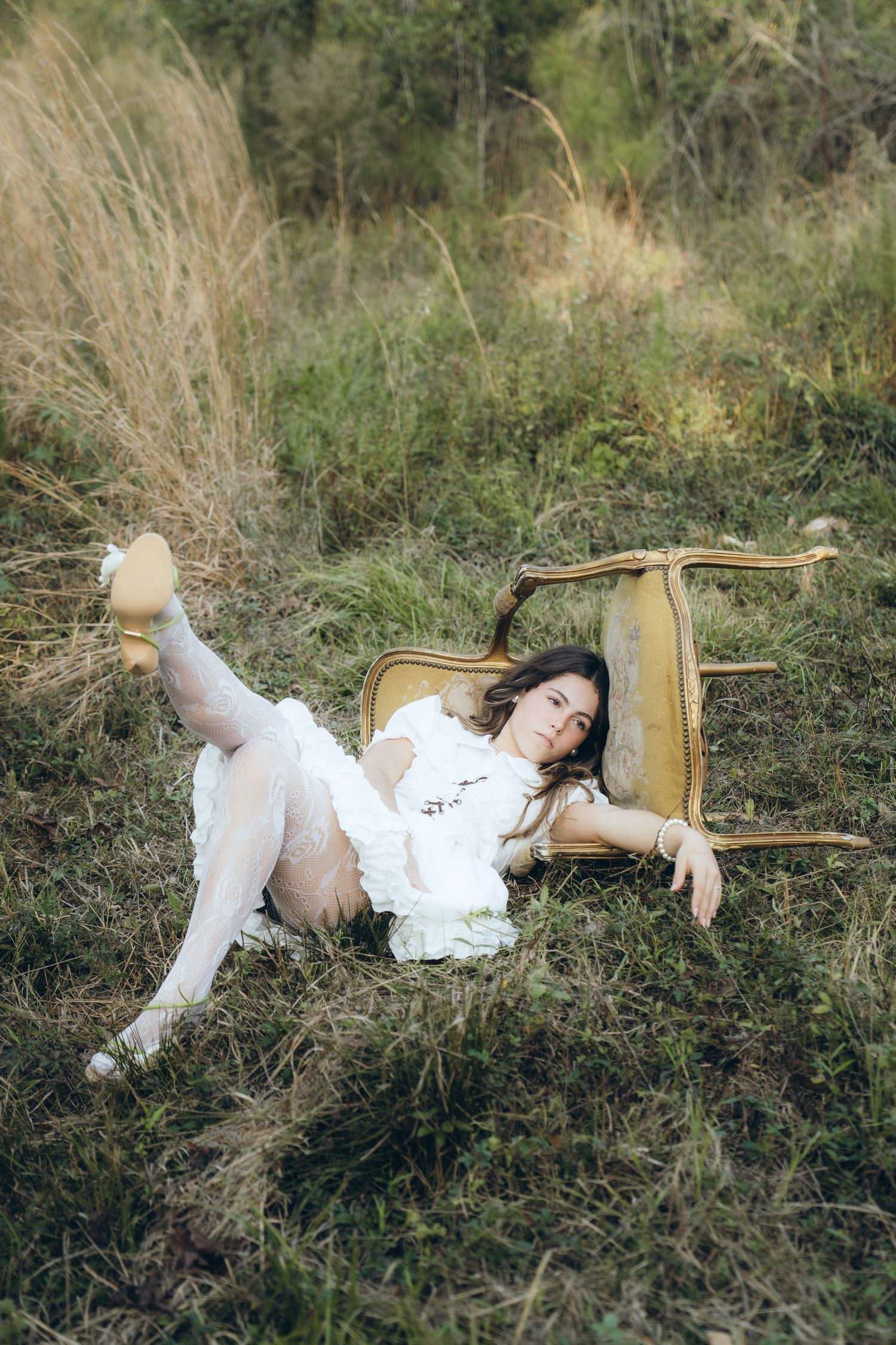 A person in a white dress lies peacefully in tall grass next to a vintage wooden chair in a dreamy outdoor setting.