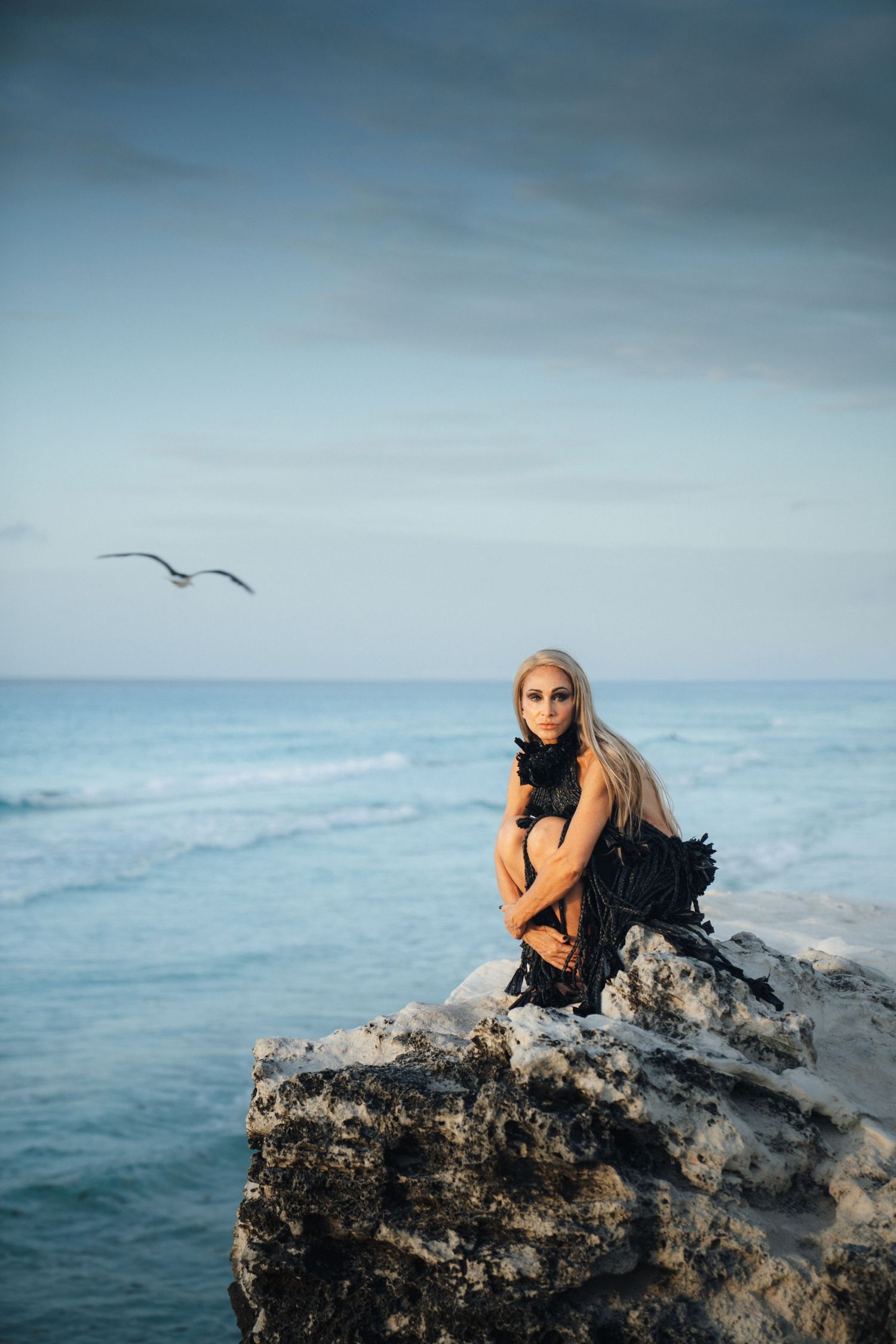 Person in contemplative pose sits atop coastal rocks with ocean waves and moody cloudy sky in background.
