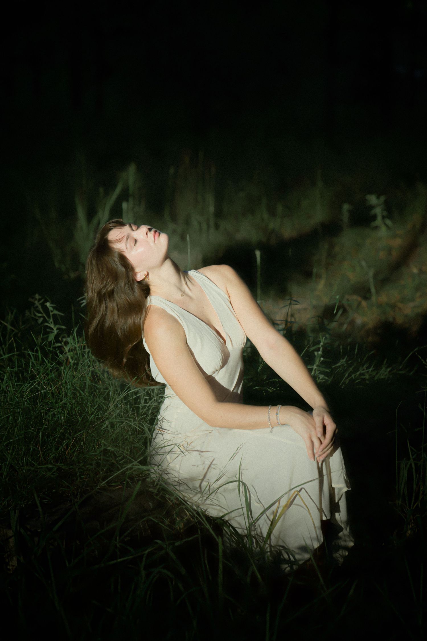 A woman in a white dress reclines in tall grass at night with dramatic lighting creating a mystical atmosphere.