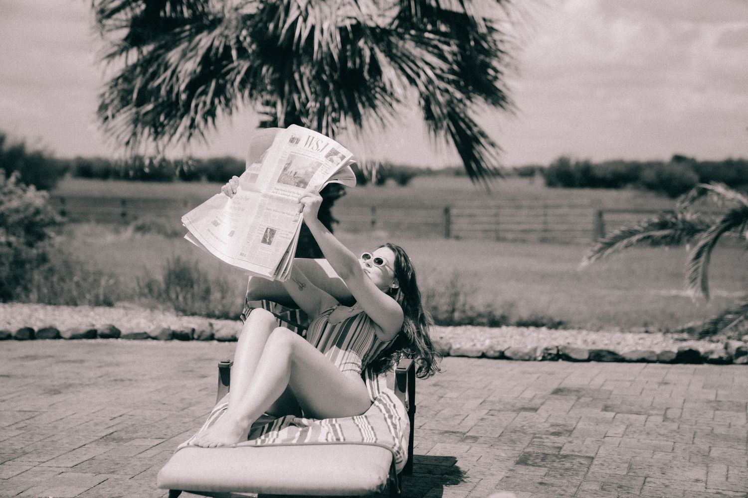 Black and white stylized photo of someone lounging casually on a deckchair with a newspaper under palm trees.