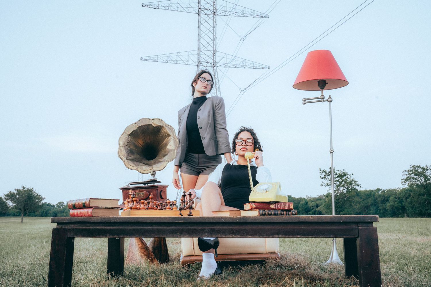 Two people pose with vintage furniture and a gramophone on a wooden table in an outdoor field setting beneath power lines.