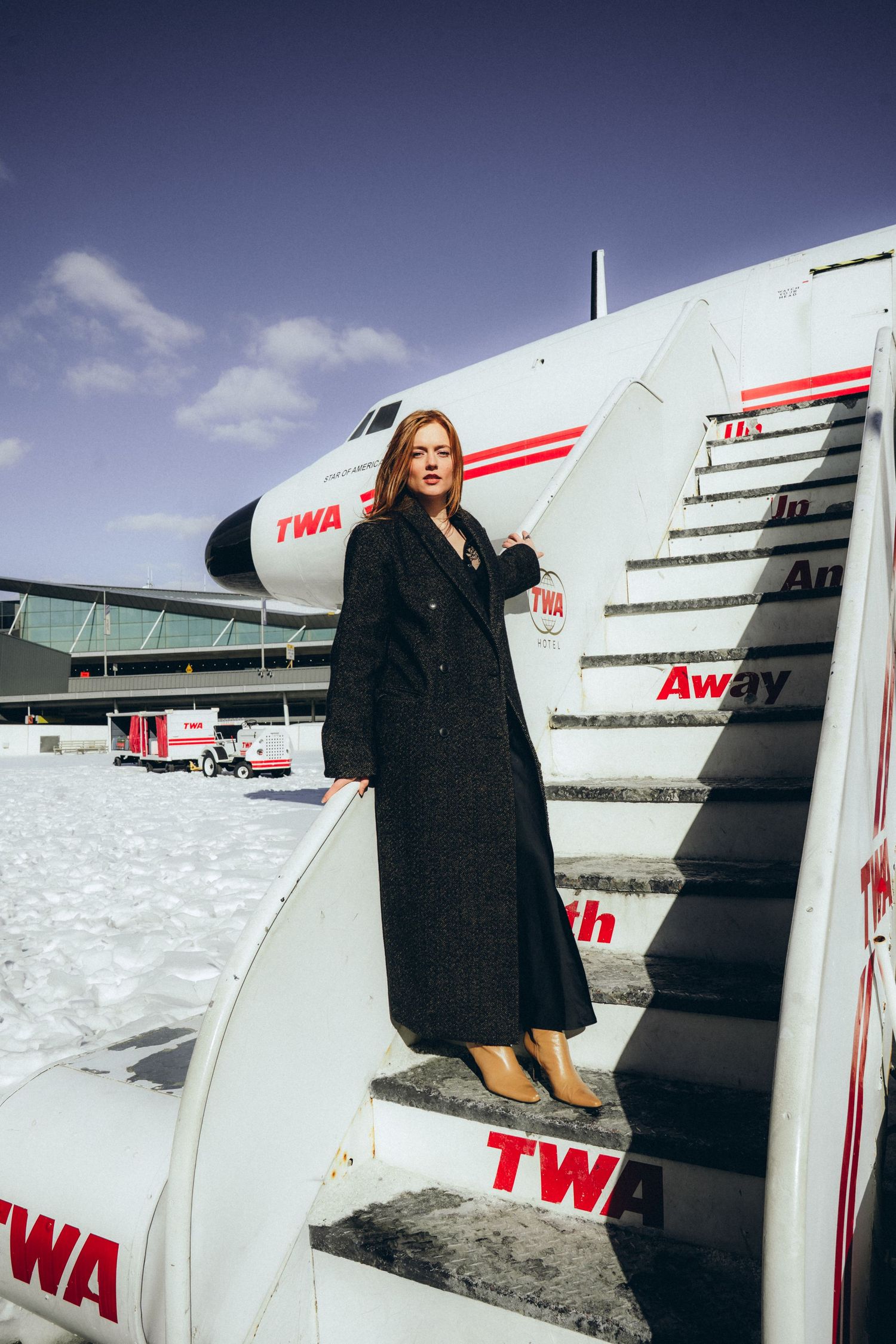 A woman in a black coat poses near vintage TWA airplane stairs against a blue sky backdrop.