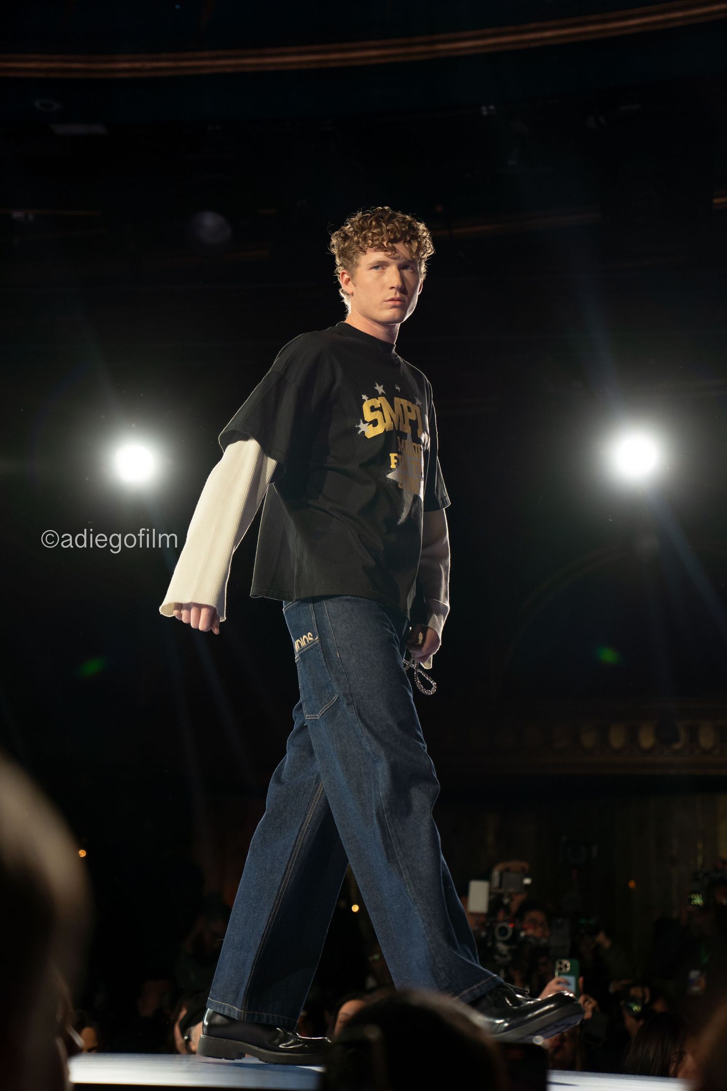 A model walks down a dark runway wearing a black t-shirt and jeans during a fashion show.