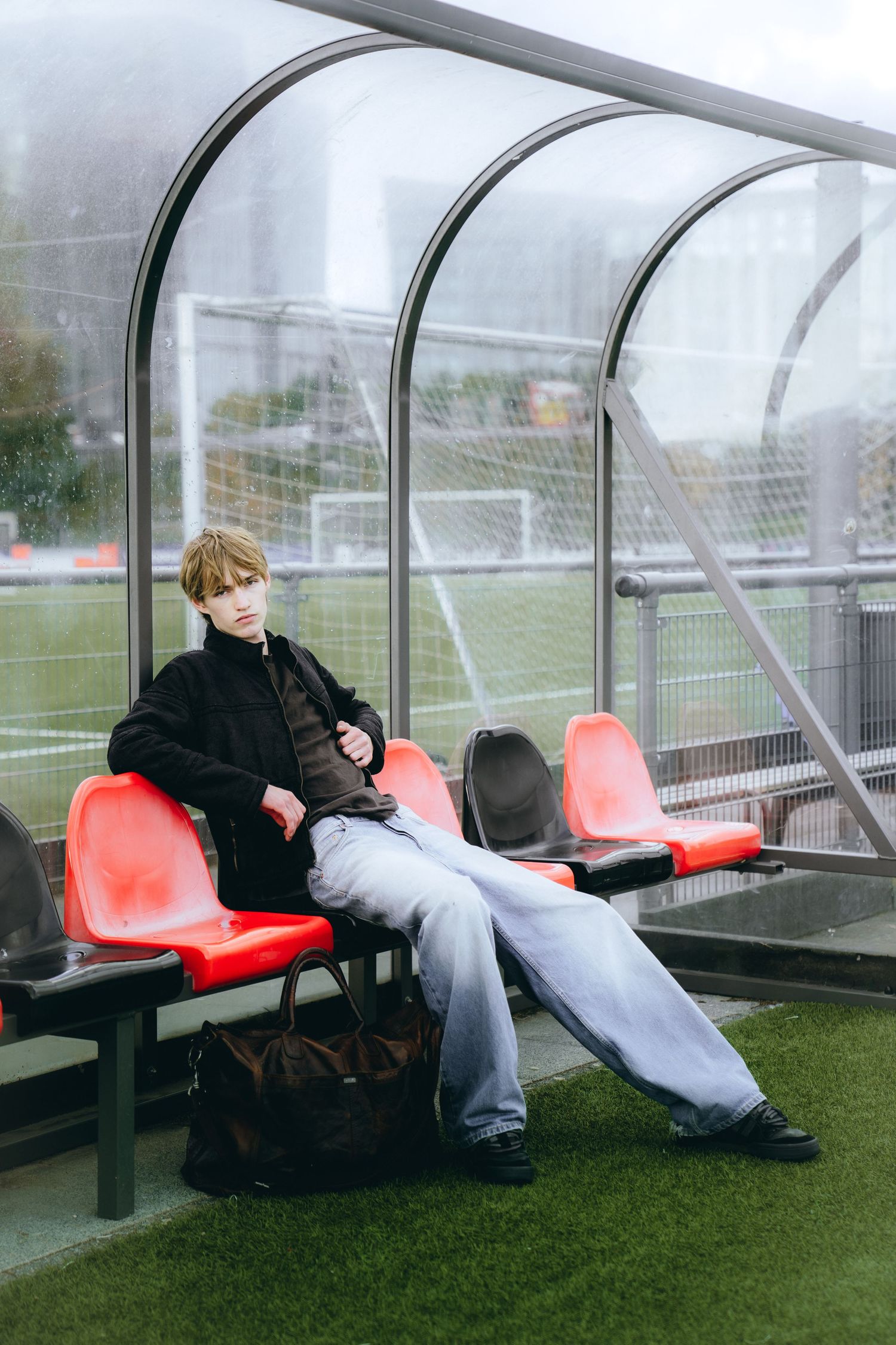Two figures rest on red bench seats in a curved glass shelter on a foggy day at a sports field.