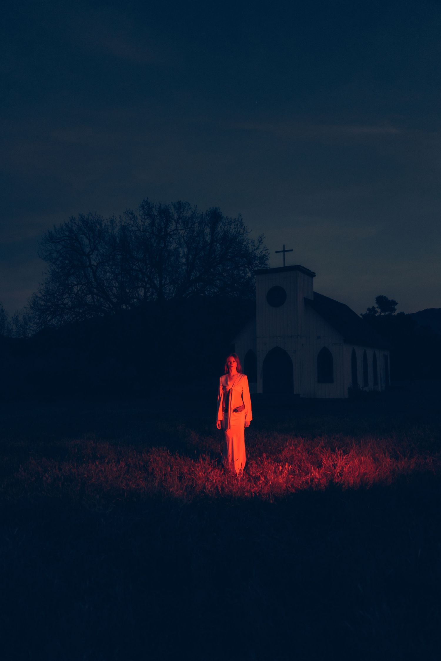 A glowing red figure stands ominously in a dark field against a moody night sky creating an eerie silhouette.