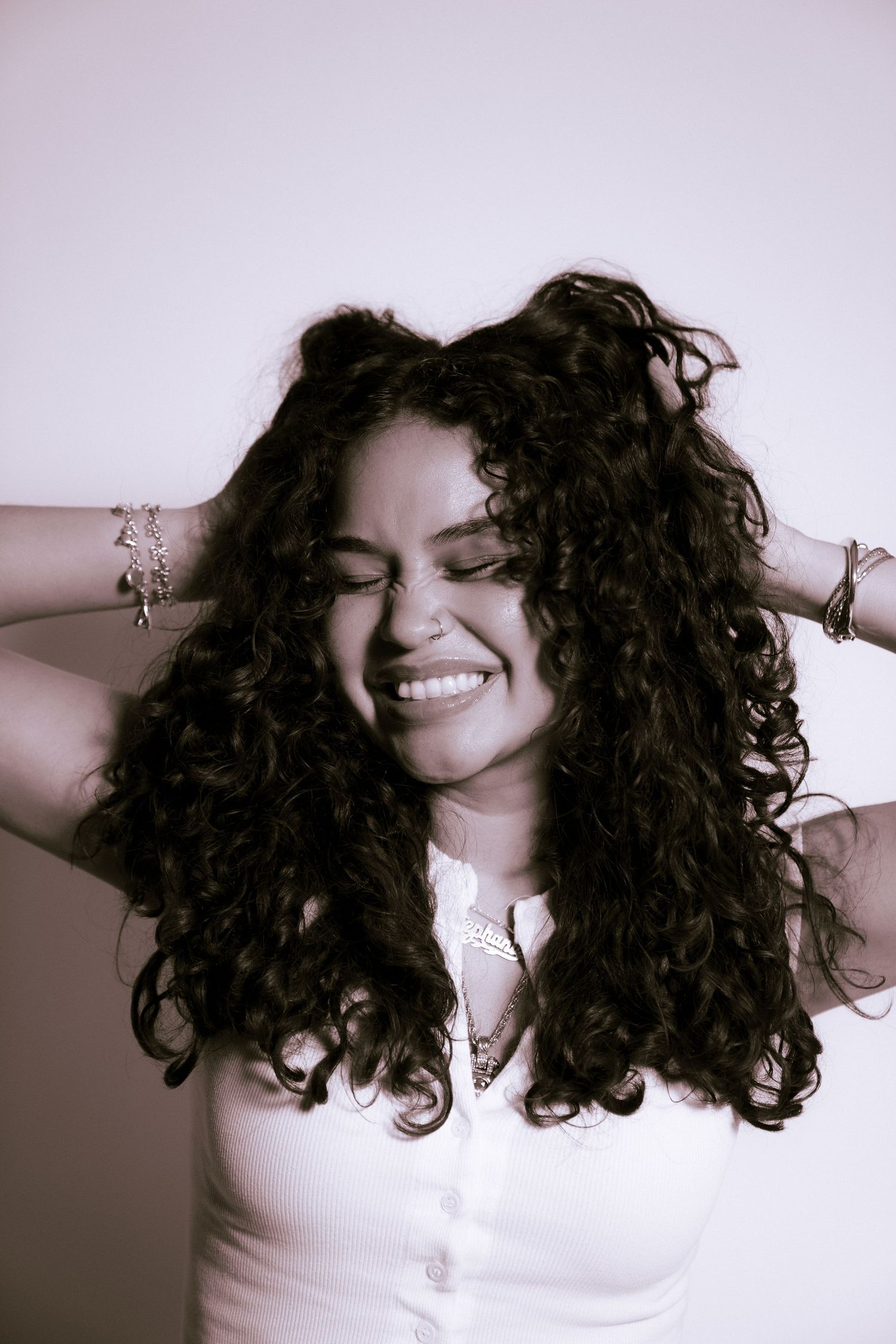 Person with long dark curly hair wearing a white top smiles joyfully while running fingers through their hair.