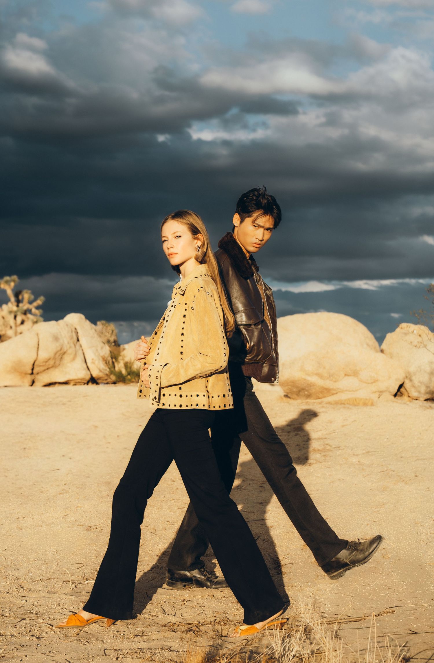 Two people walk across desert sand against dramatic stormy clouds in a panoramic series of motion shots.