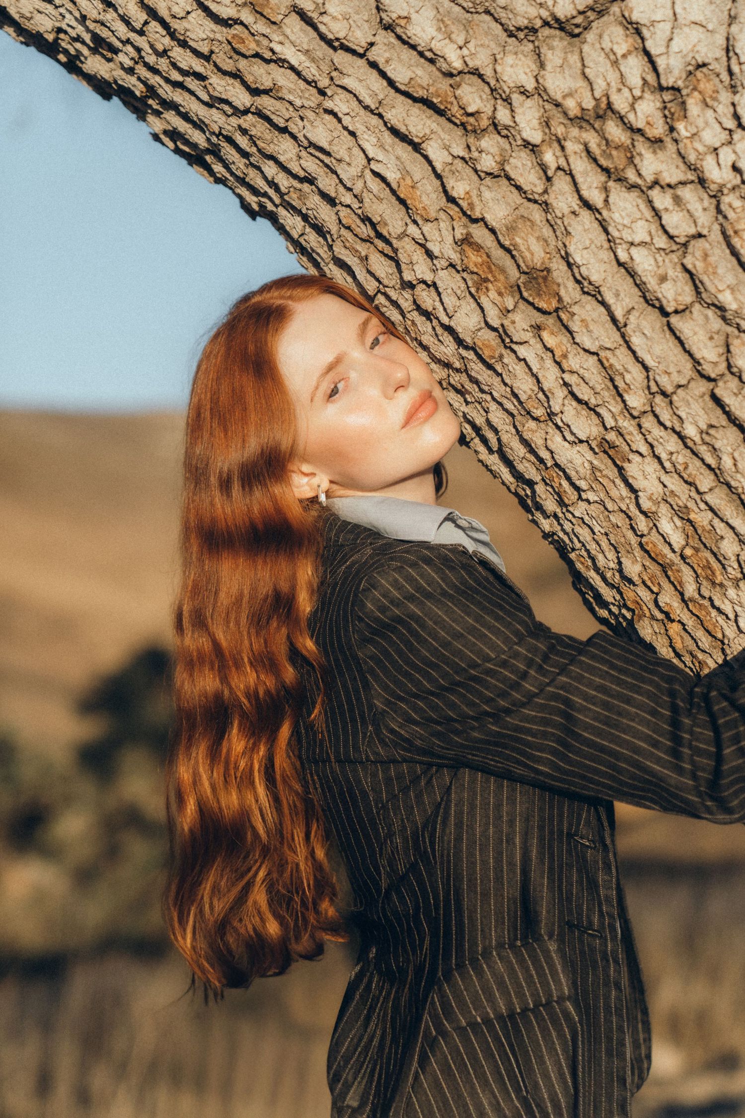 Person with long red hair wearing a black striped sweater leans against a tree trunk in an outdoor setting.