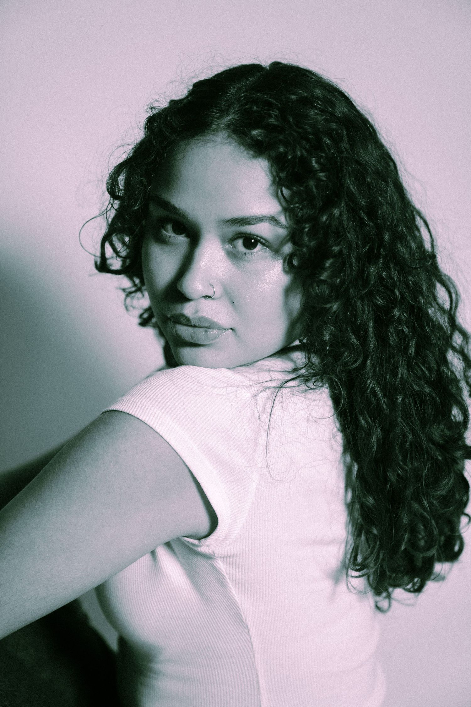 Black and white moody portrait with curly dark hair and white sleeveless top against pink background.