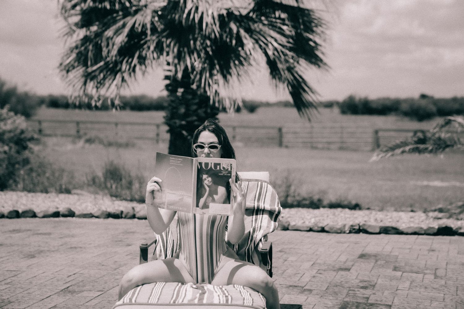 Vintage black and white photograph of someone sitting in a striped chair outdoors with a palm tree in the background.