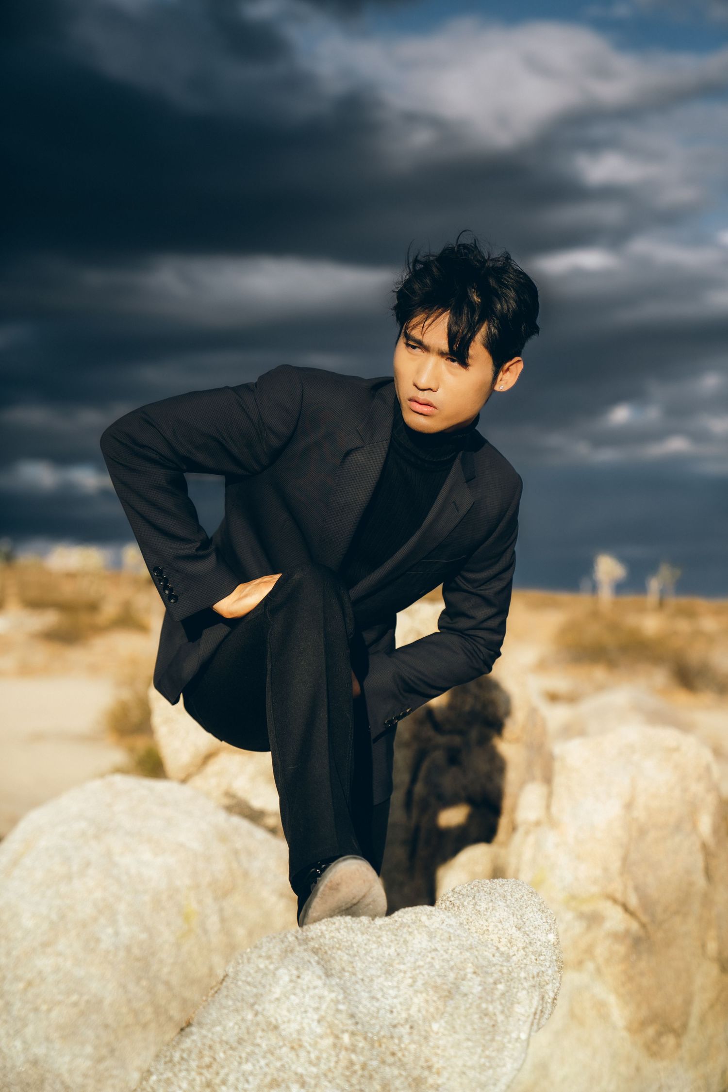 Young Asian man in black turtleneck suit poses on desert rocks under dramatic stormy sky.