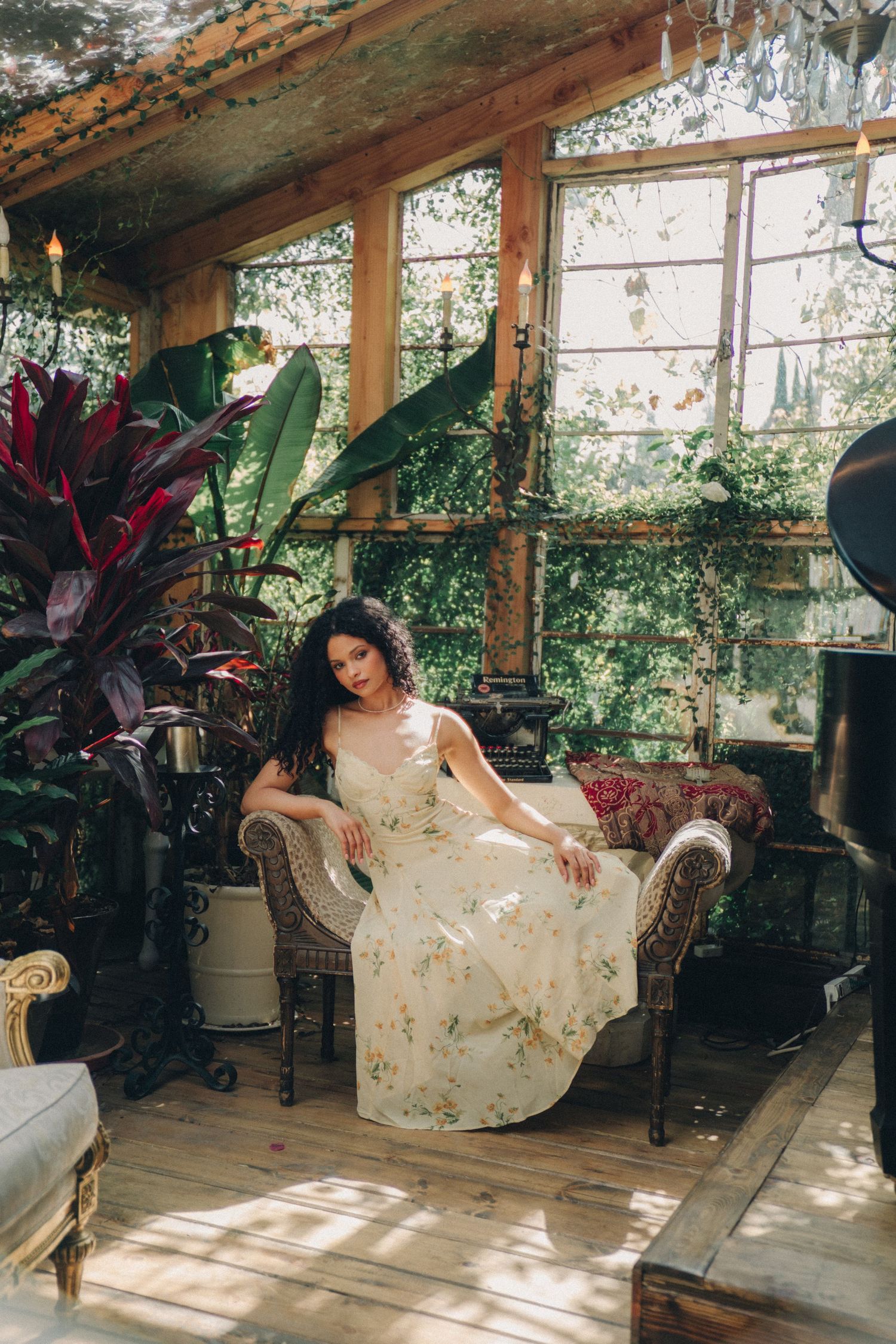 Woman in floral maxi dress seated in a wicker chair surrounded by lush tropical plants in a sunlit bohemian greenhouse.