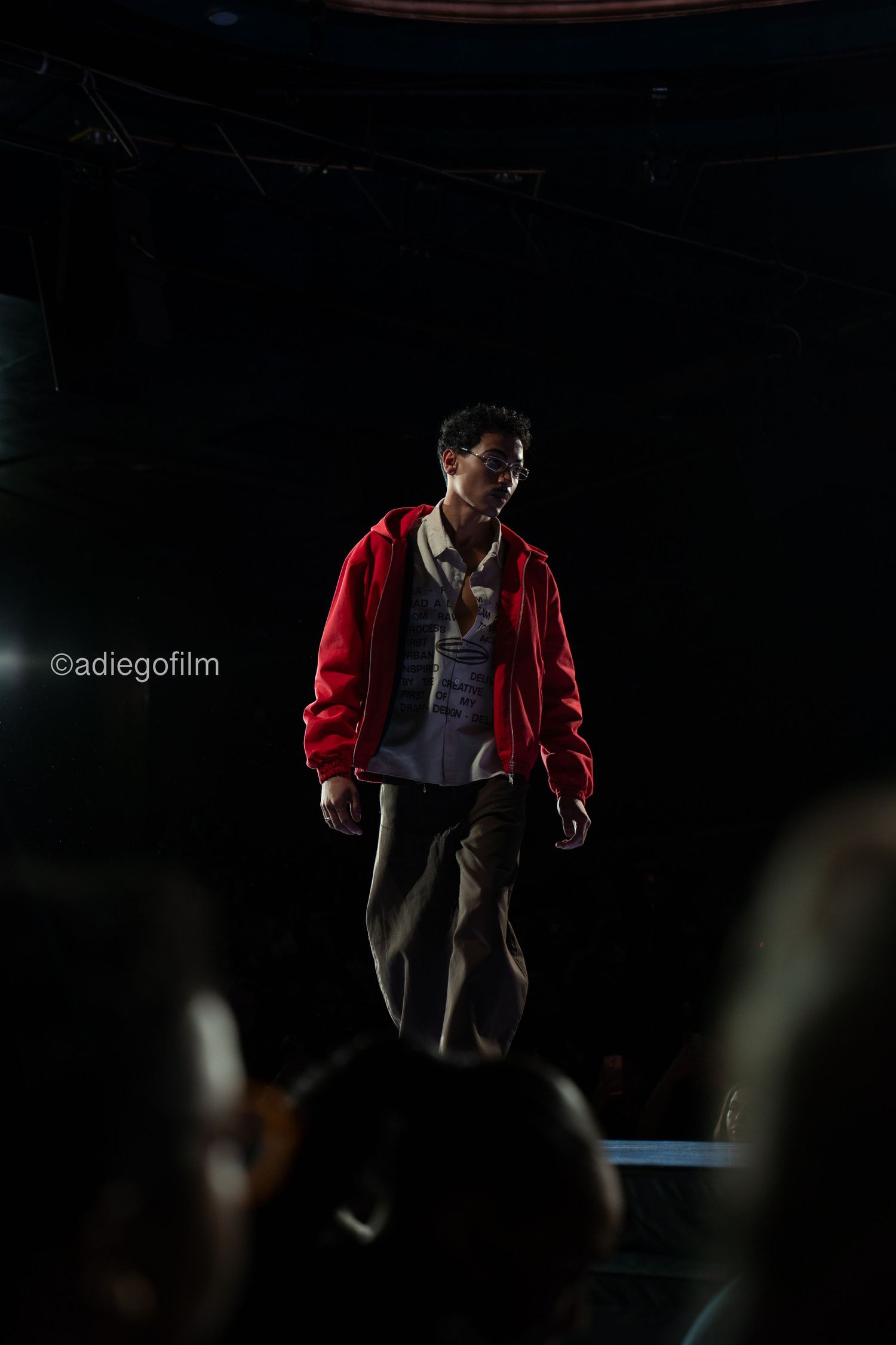 Figure in a red jacket performing on a dimly lit stage with theatrical backlighting creating a moody atmosphere.