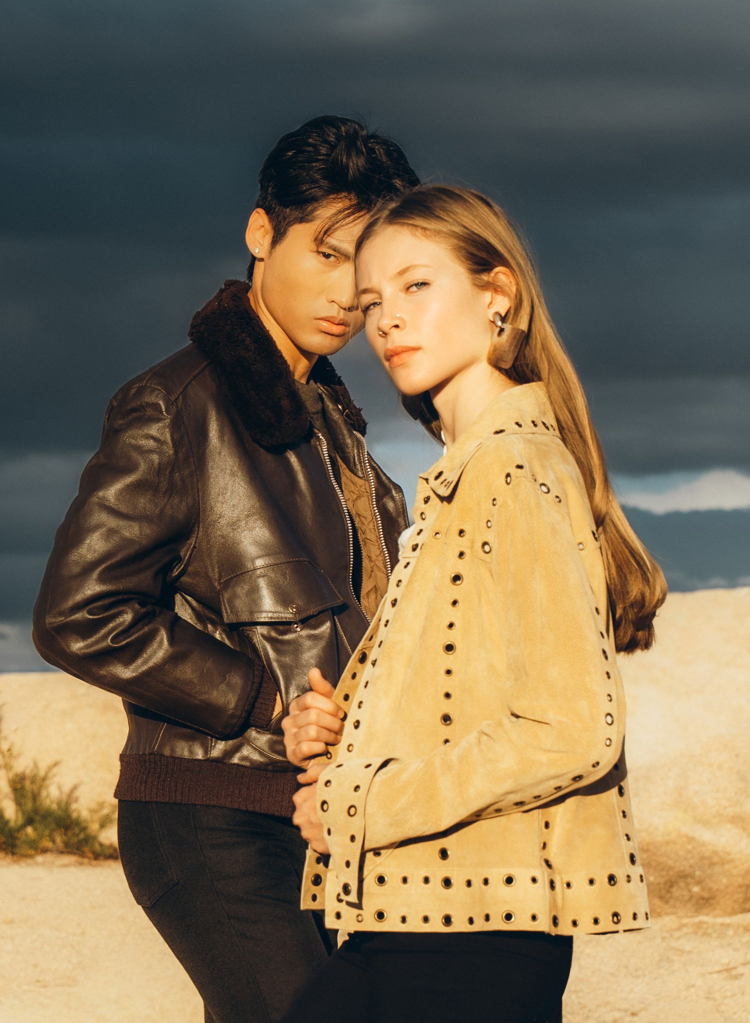 Romantic couple in brown leather and beige outfits embrace against desert backdrop at golden hour.