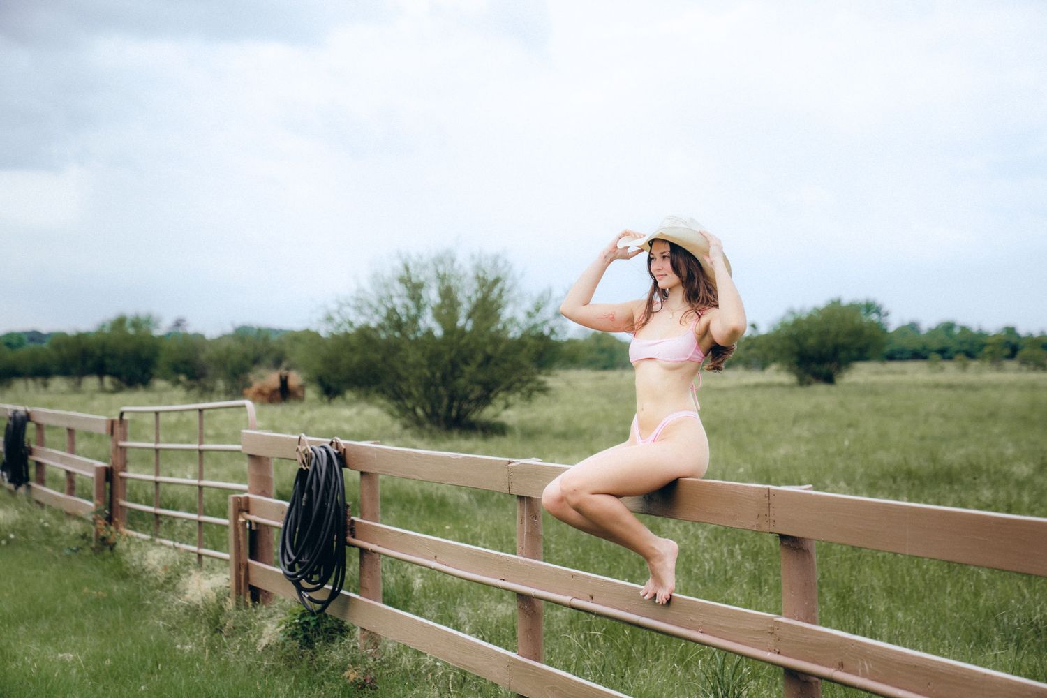 Someone in a sundress and hat sits on a wooden fence overlooking a pastoral green field on a summer day.