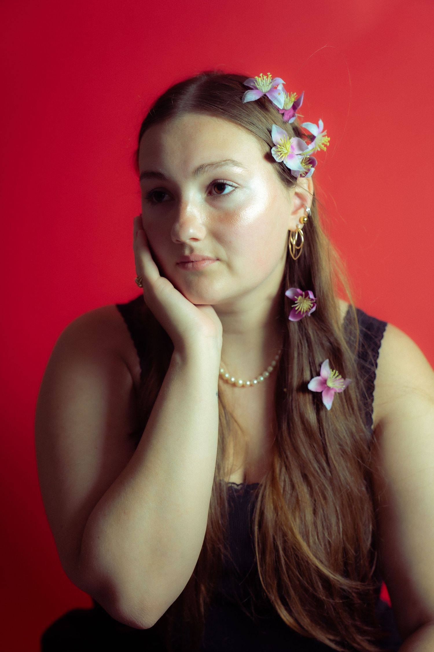 A person in front of red background wearing flowers in their hair poses with hand on chin in a contemplative manner.