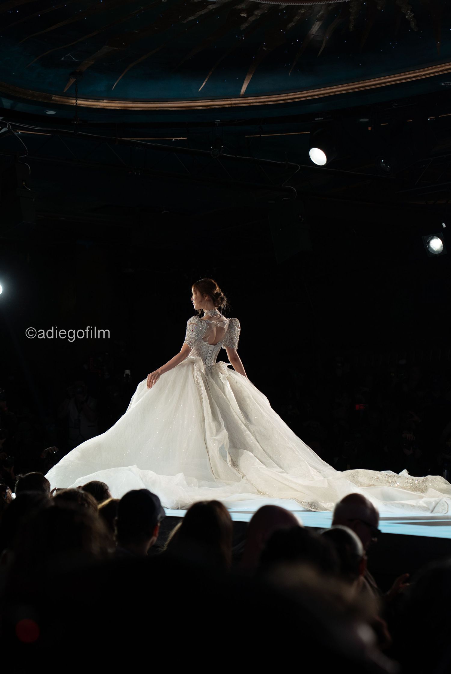 A bride in a flowing white wedding gown twirls elegantly on a dark stage during a bridal fashion show.