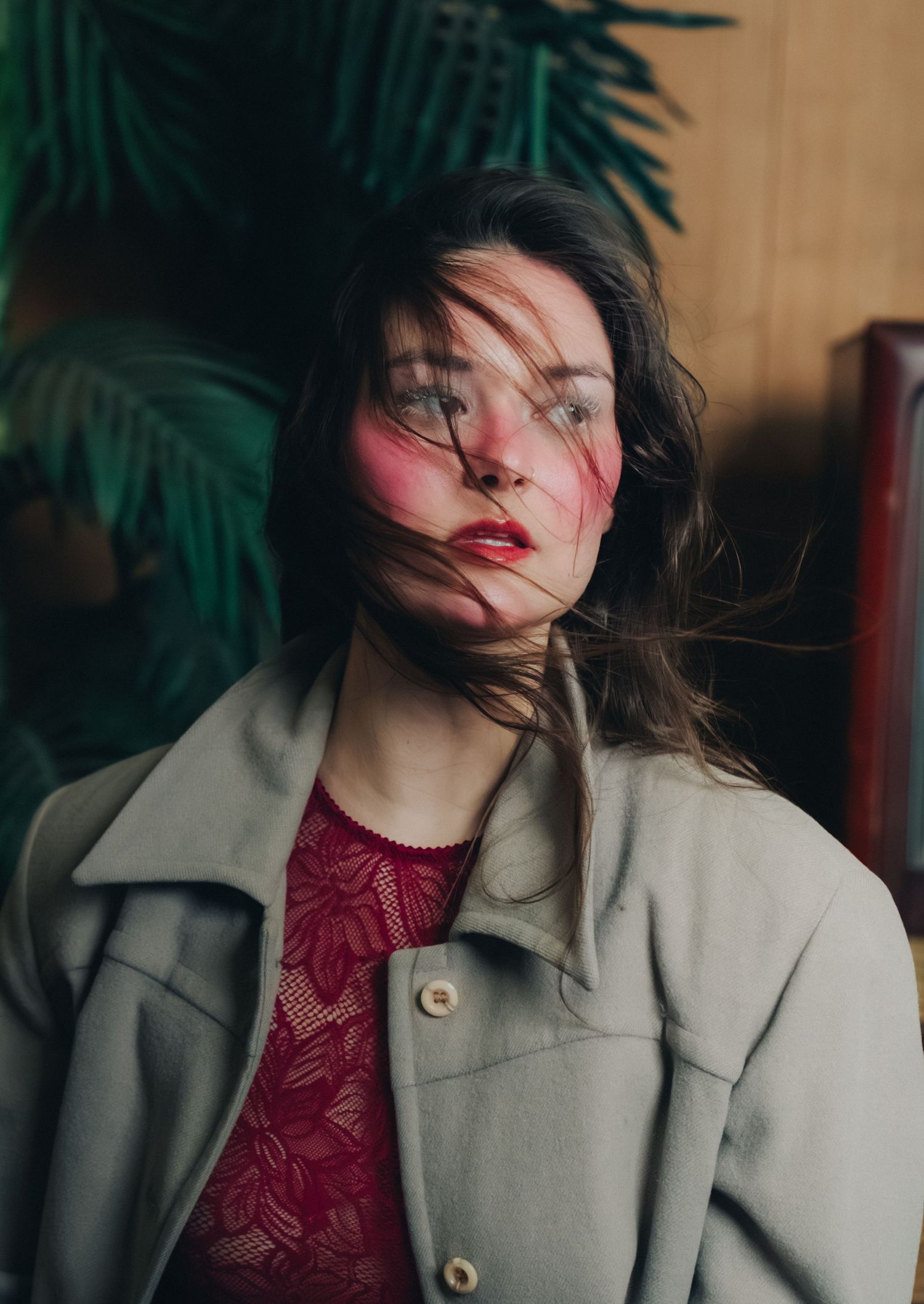 Fashion portrait of person in beige coat and red top with dark hair against tropical plant background.