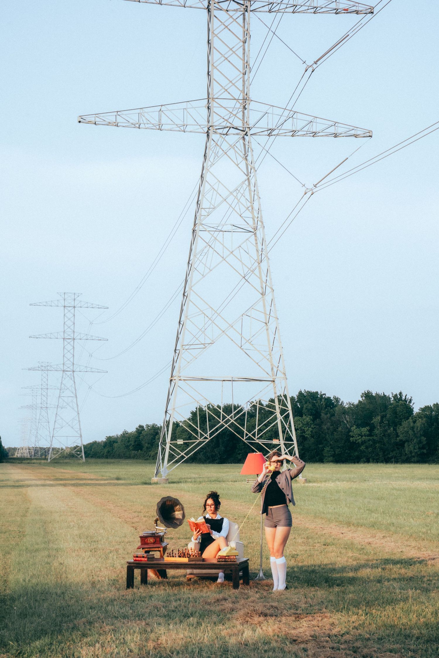 Group relaxing on wooden bench under large electrical transmission tower in grassy field on sunny day.